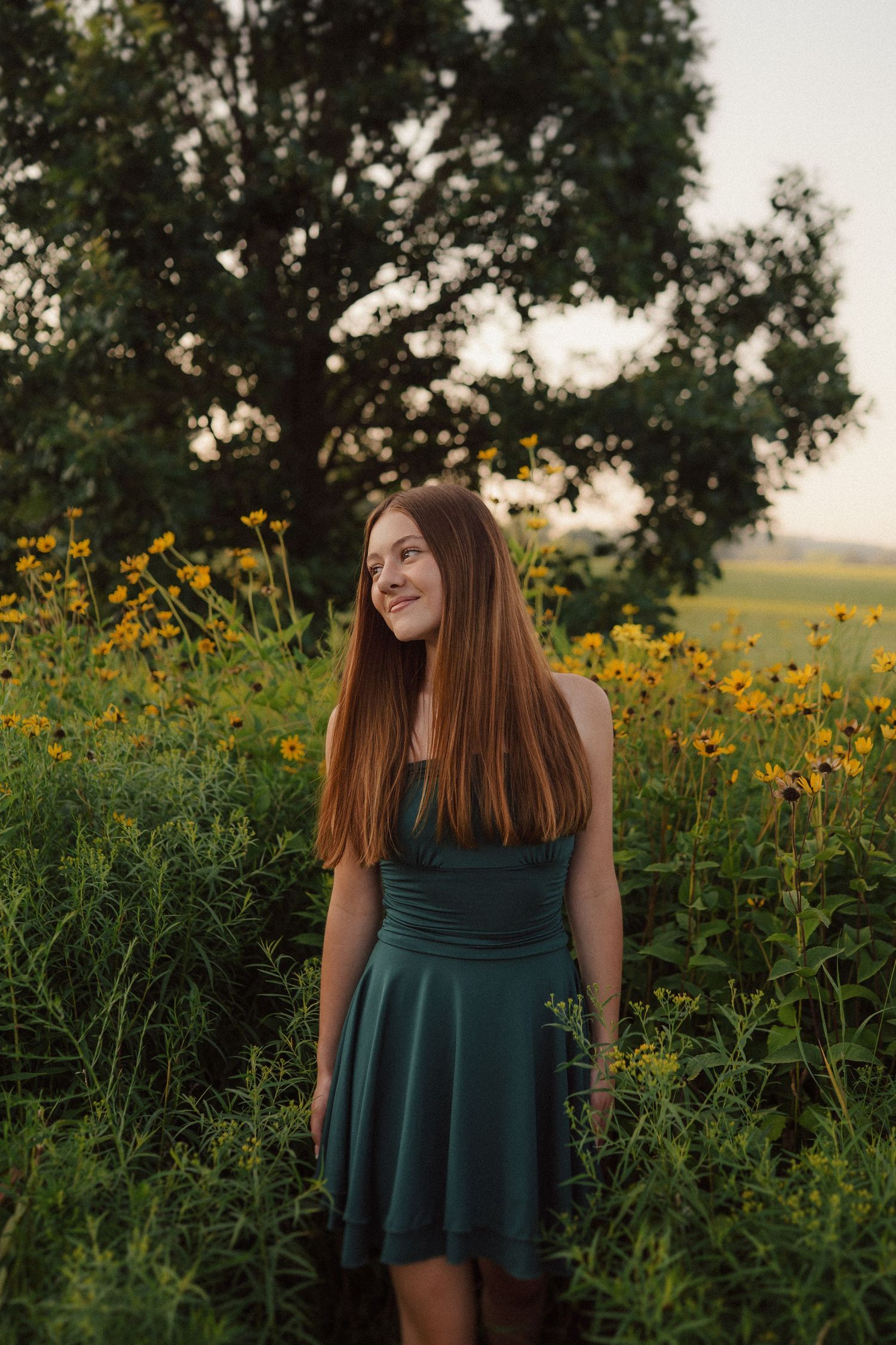 A person in a dark green dress stands among blooming yellow wildflowers at sunset in a rural field.