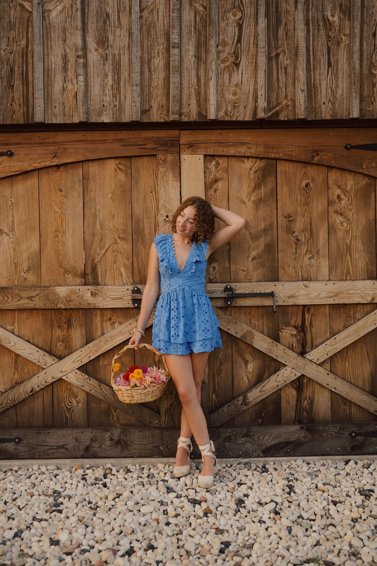 Figure in a blue dress poses playfully against rustic wooden barn wall with gravel ground underneath.