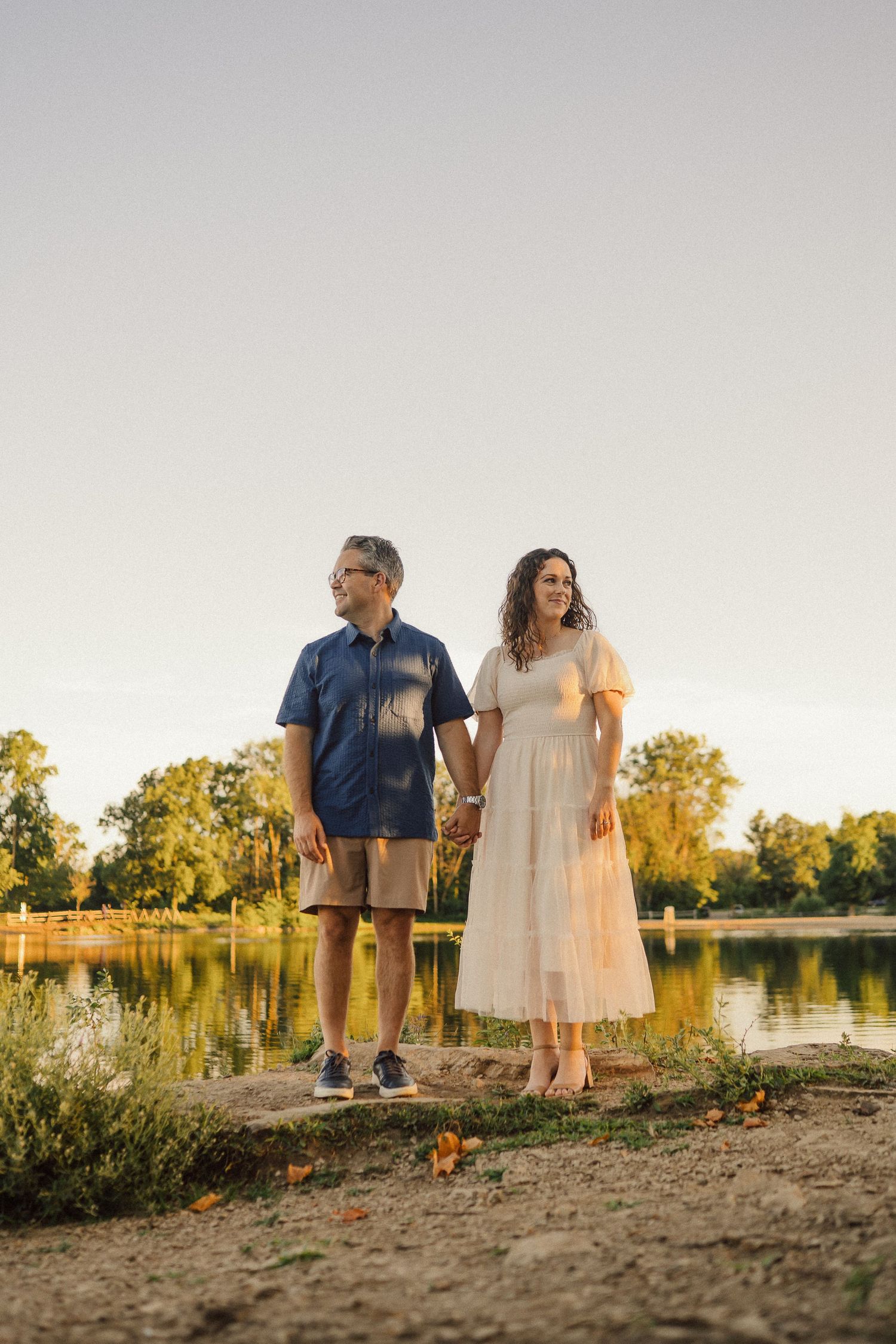 Couple standing together at golden hour by a serene lakeside in summer attire.