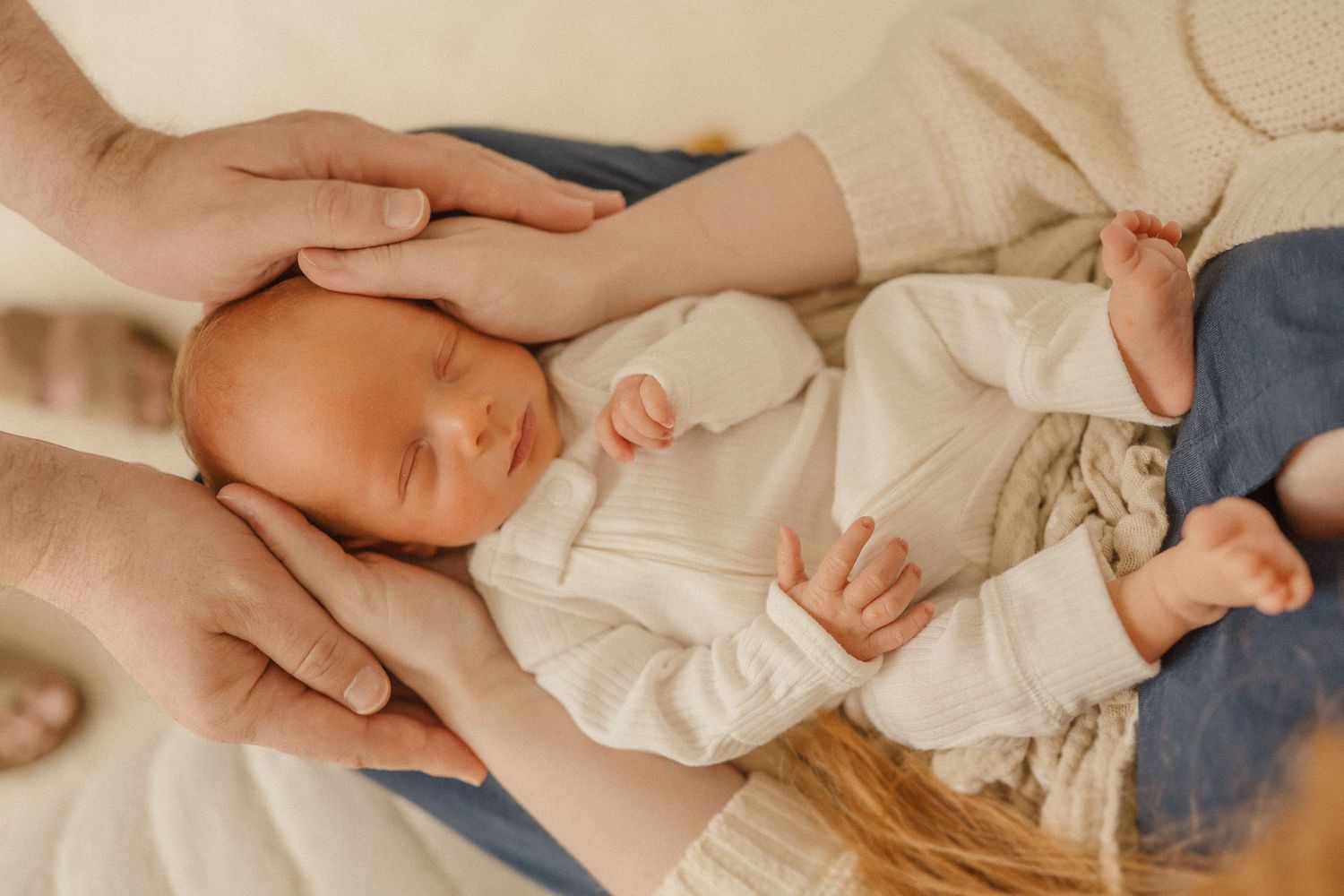 Tender moment of adult hands cradling a sleeping infant wrapped in cream colored clothing.