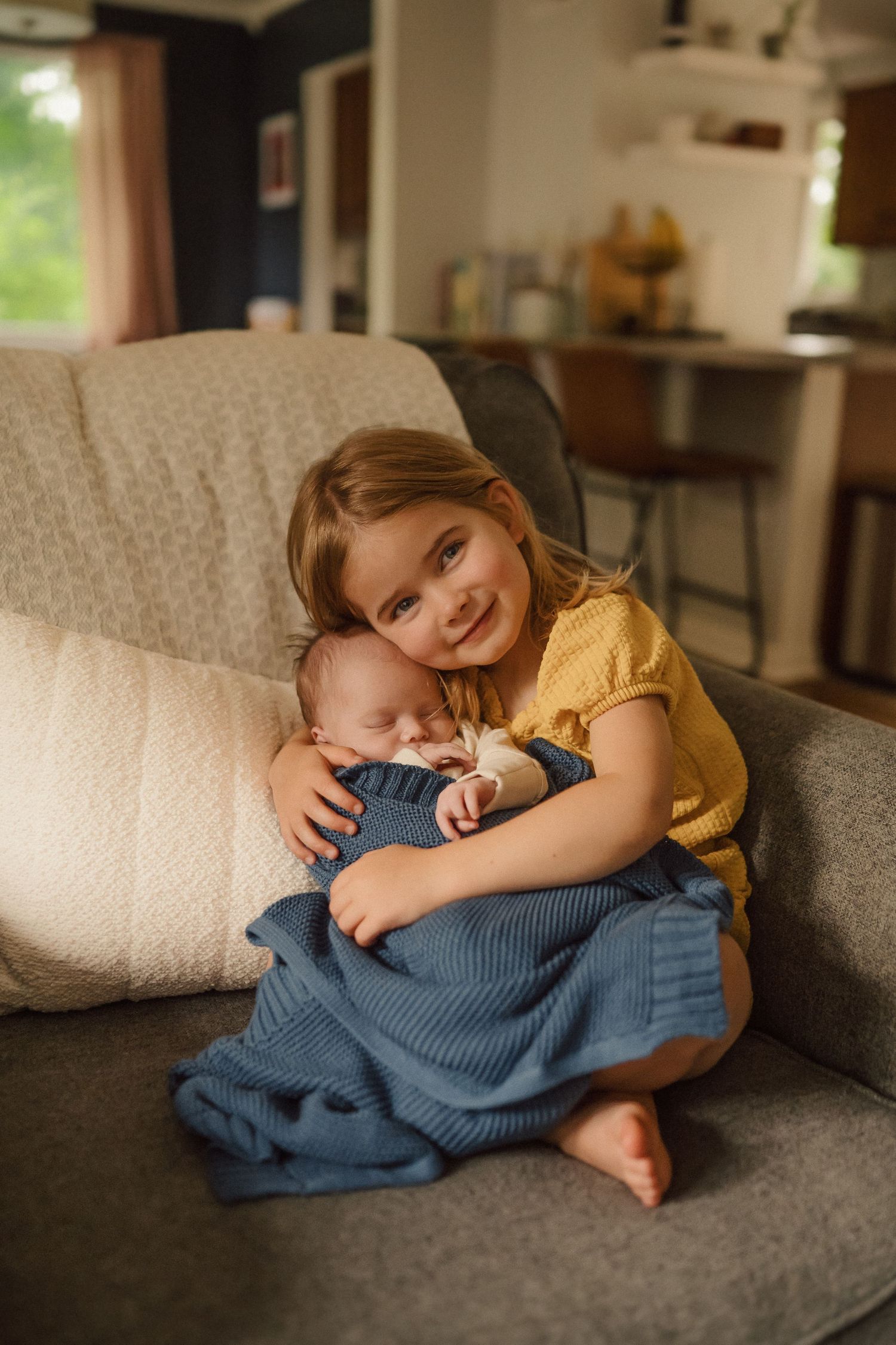 A child in a yellow shirt cuddling with an infant wrapped in blue blanket on a couch.