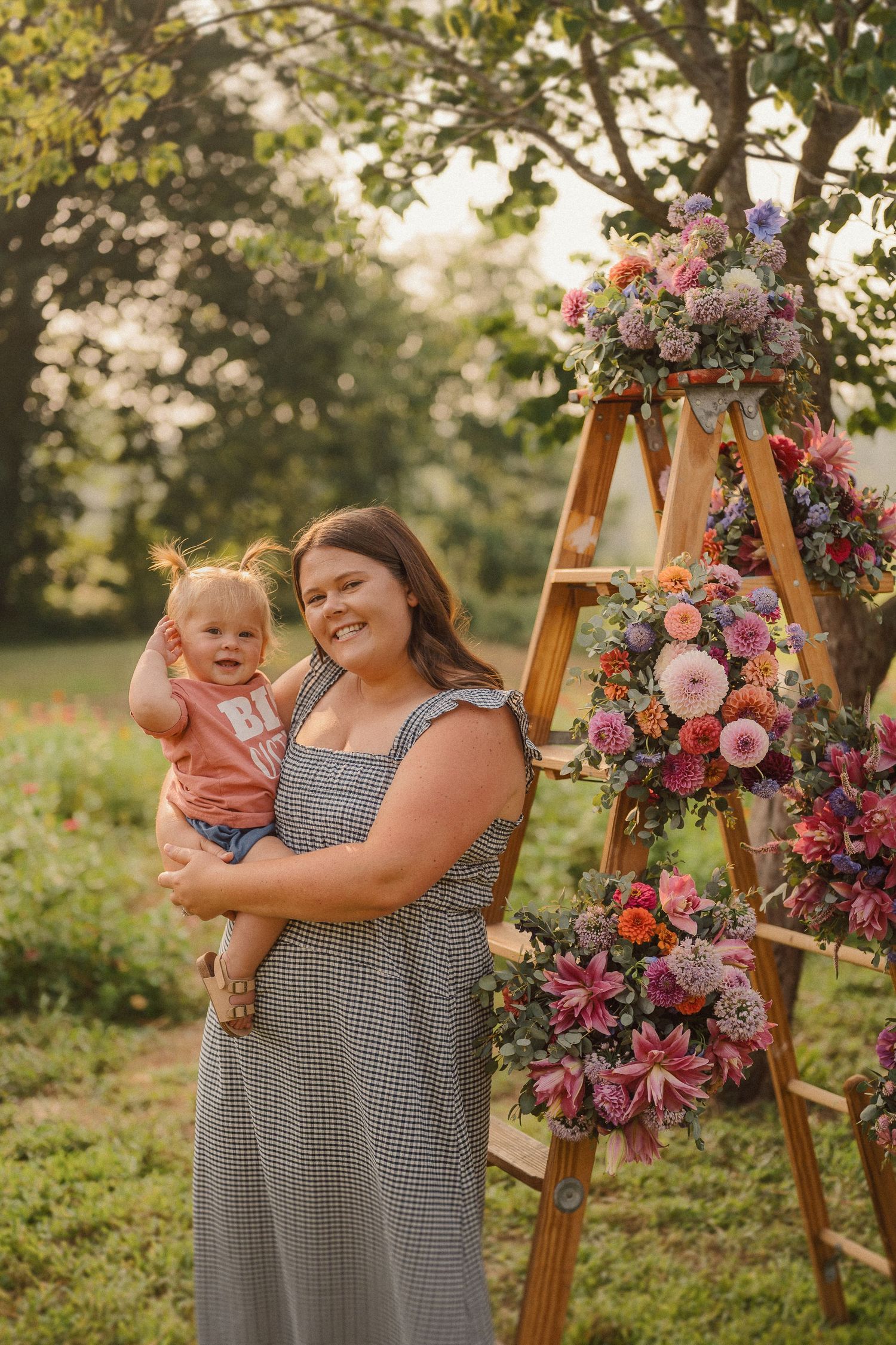 Mother and baby pose next to wooden easel decorated with pink floral arrangement in outdoor garden setting.