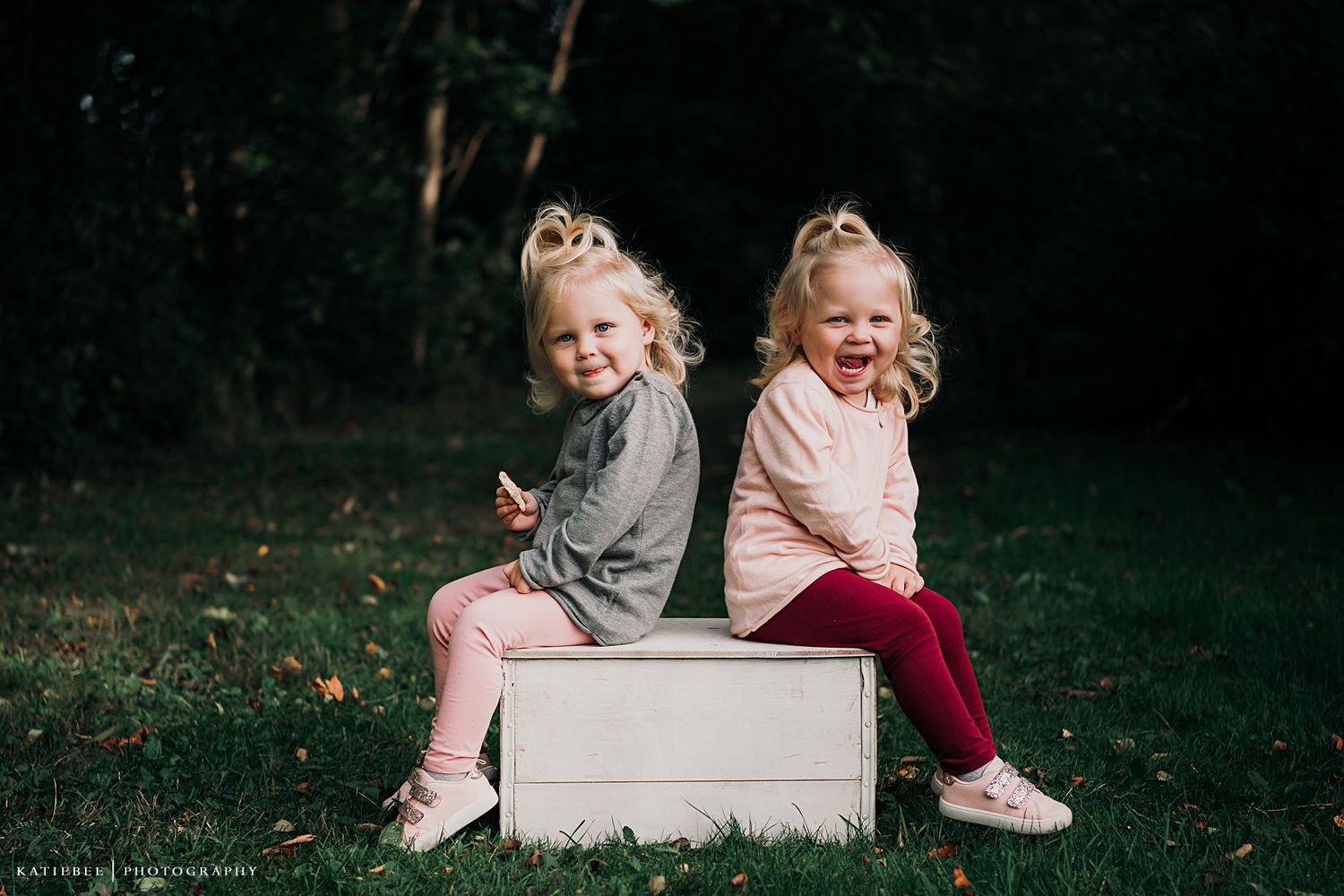 Two children sit back-to-back on a wooden box, giggling in natural outdoor setting.