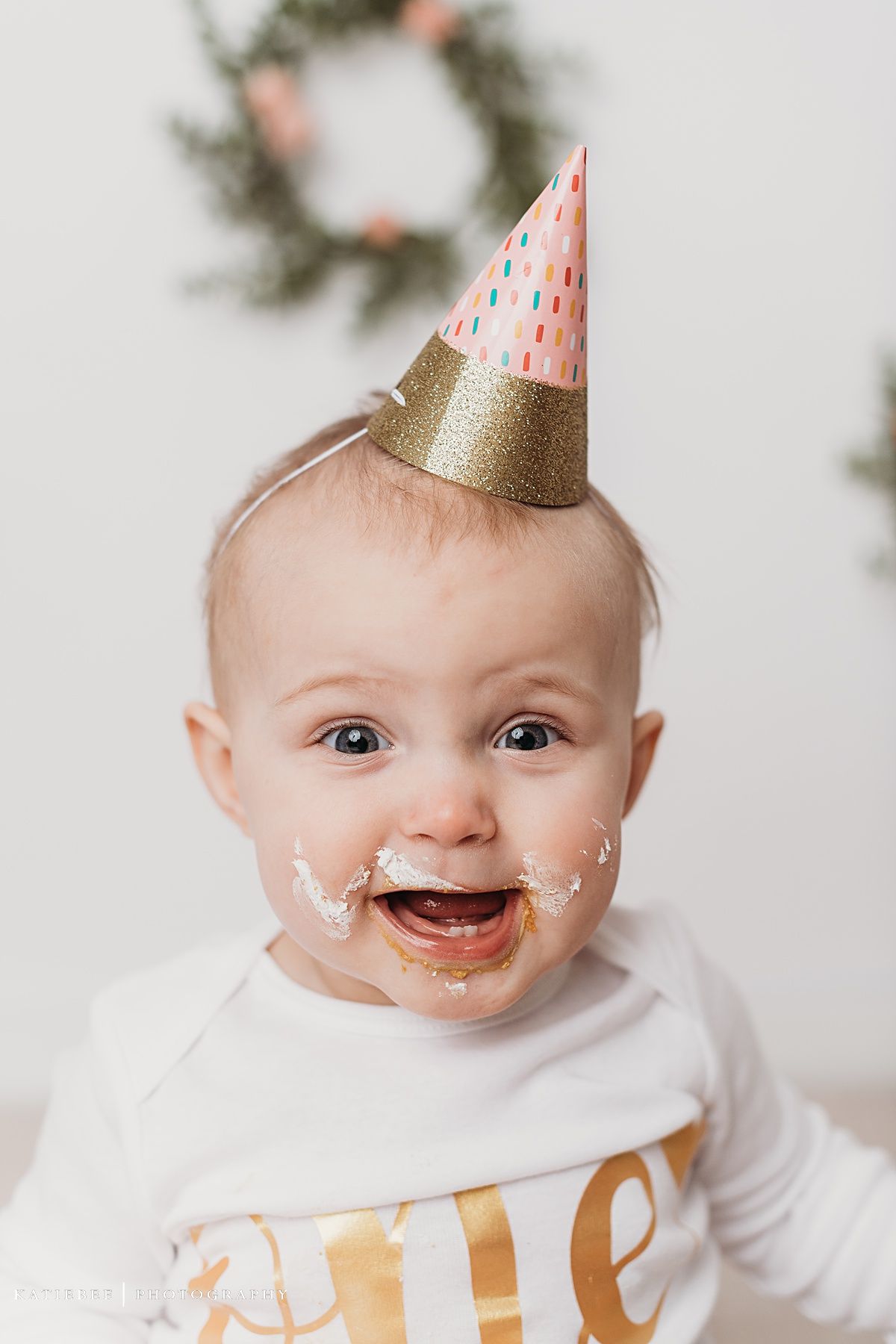 Baby wearing party hat laughing with cake frosting smeared on cheeks.