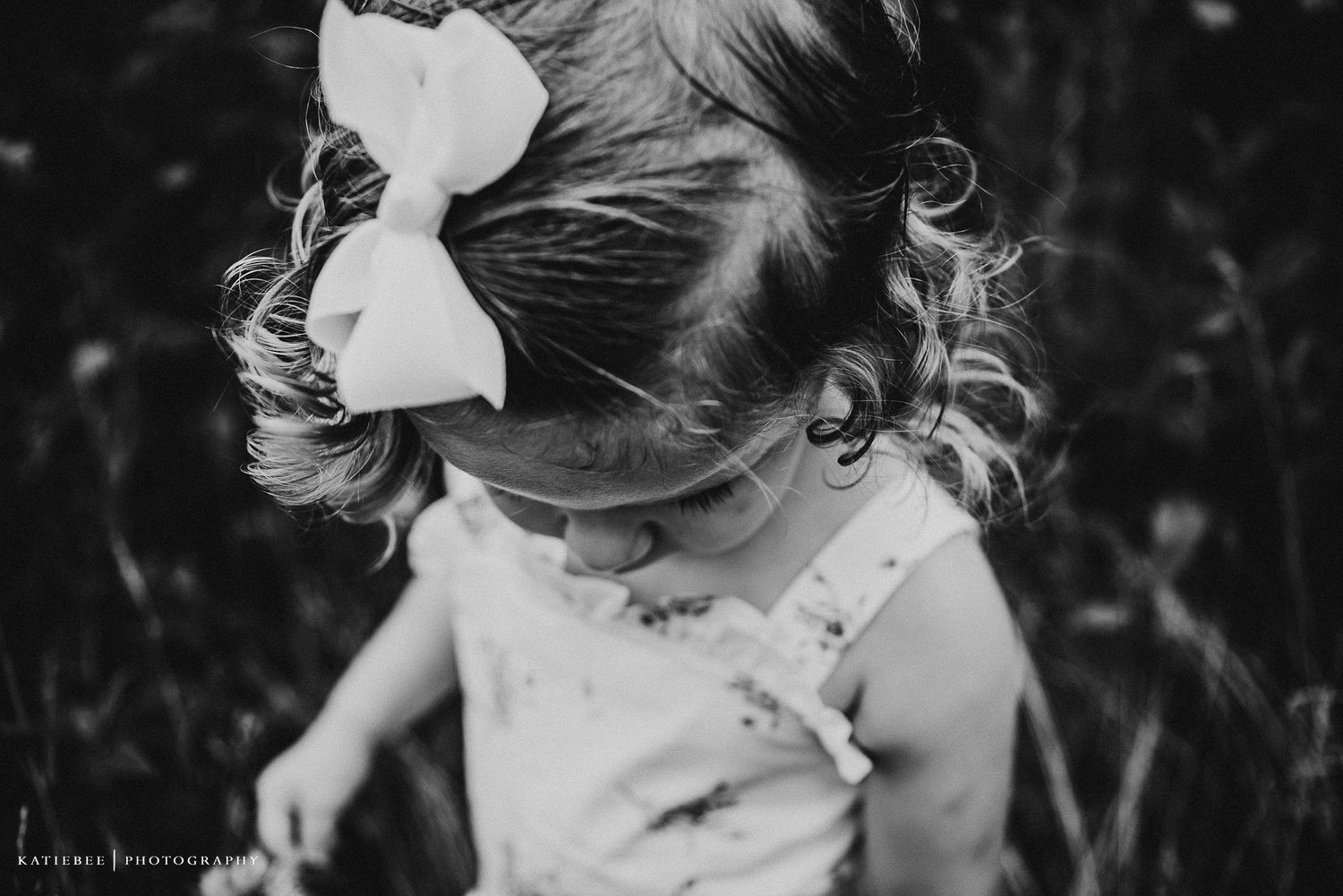 A black and white portrait of a small child wearing a white bow in their curly hair.