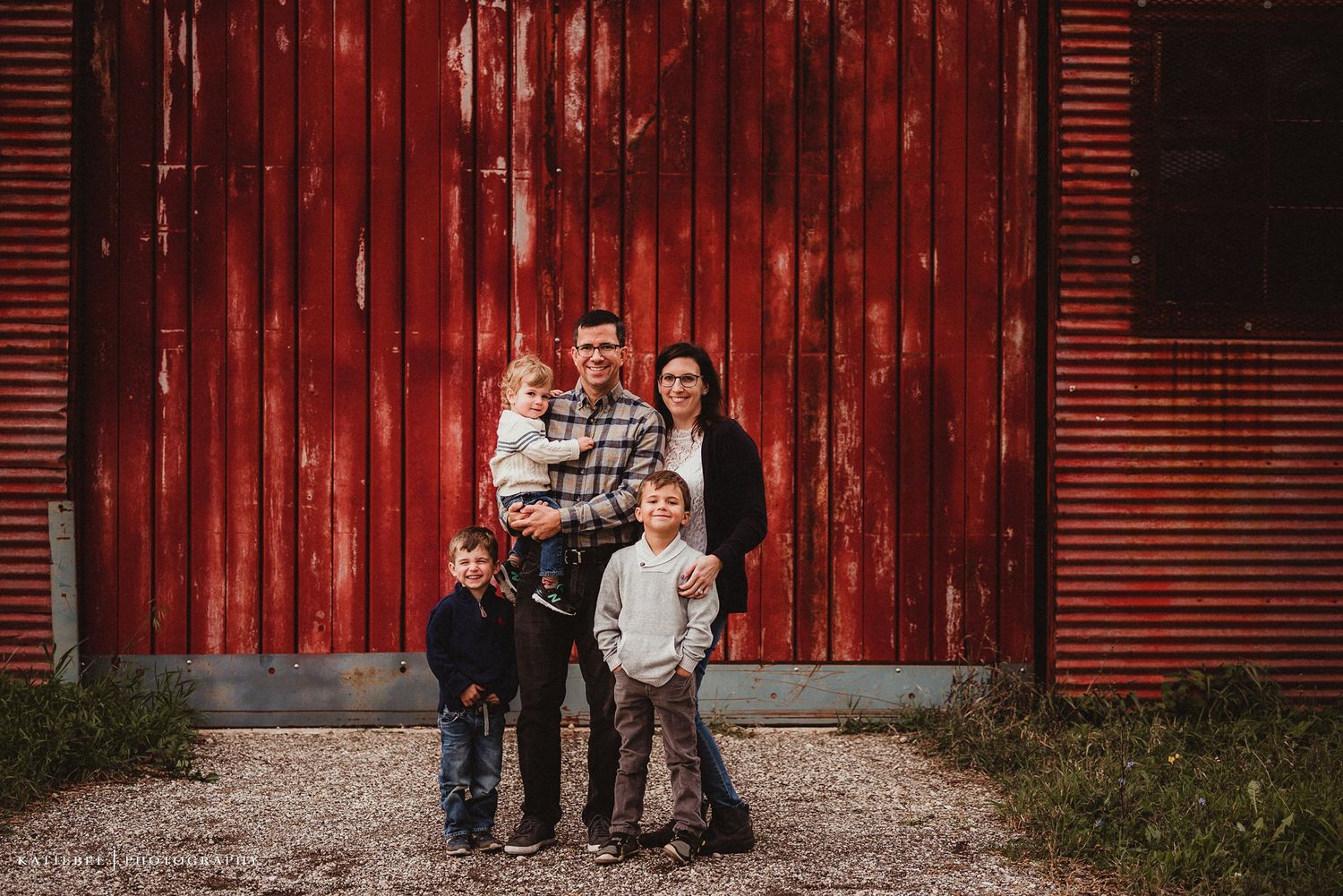 Family portrait in front of a rustic red barn door on a cloudy day.