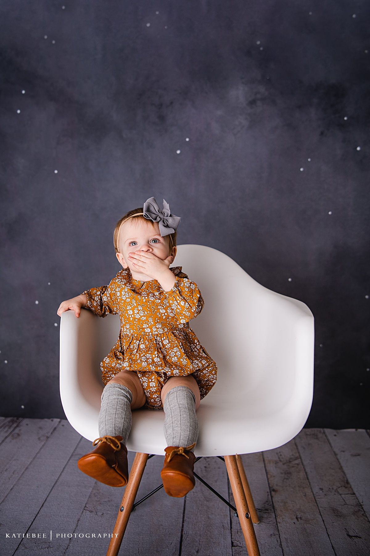 Child in mustard dress sitting in modern white chair against dark sparkly background.