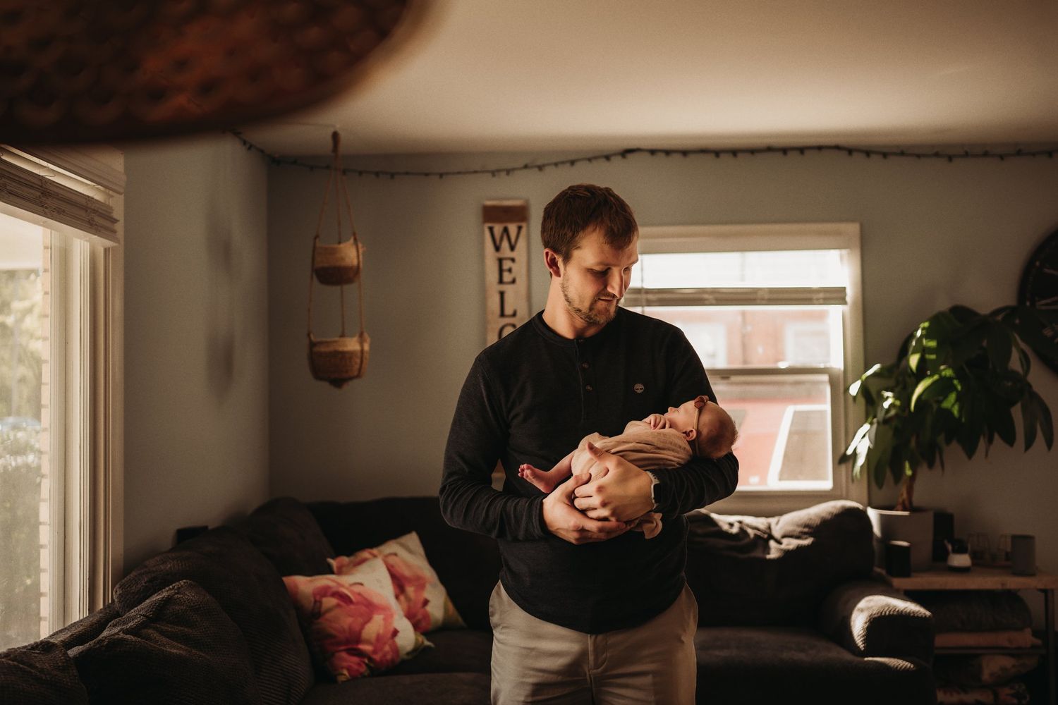 A heartwarming moment in a sunlit living room as a baby is held near large windows.
