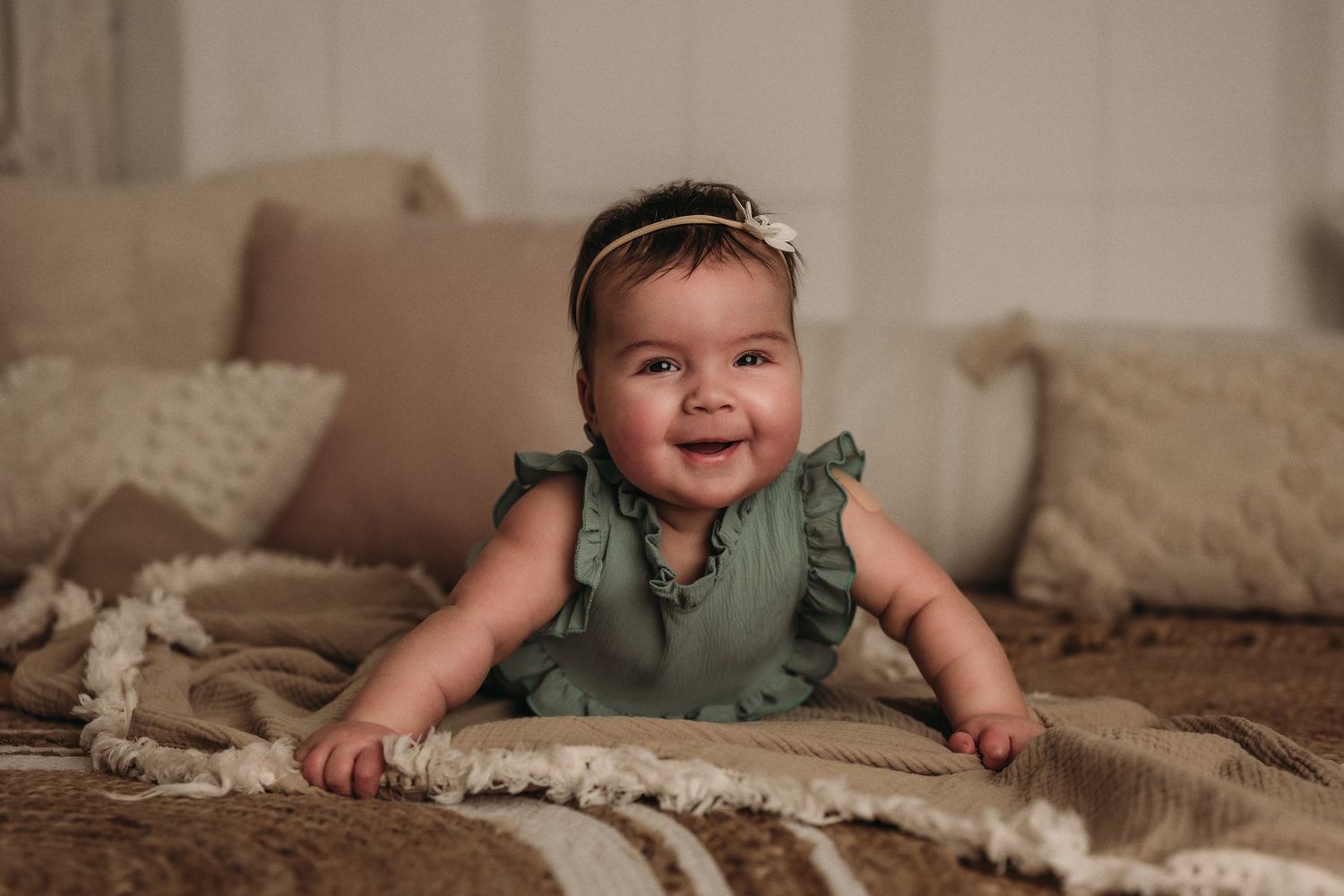 Baby in sage green ruffled outfit smiles while laying on textured beige blanket.