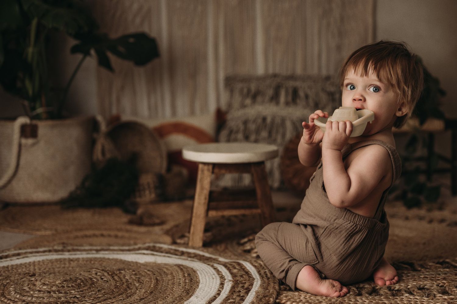 A baby in a brown romper sits on a patterned rug next to a wooden stool.