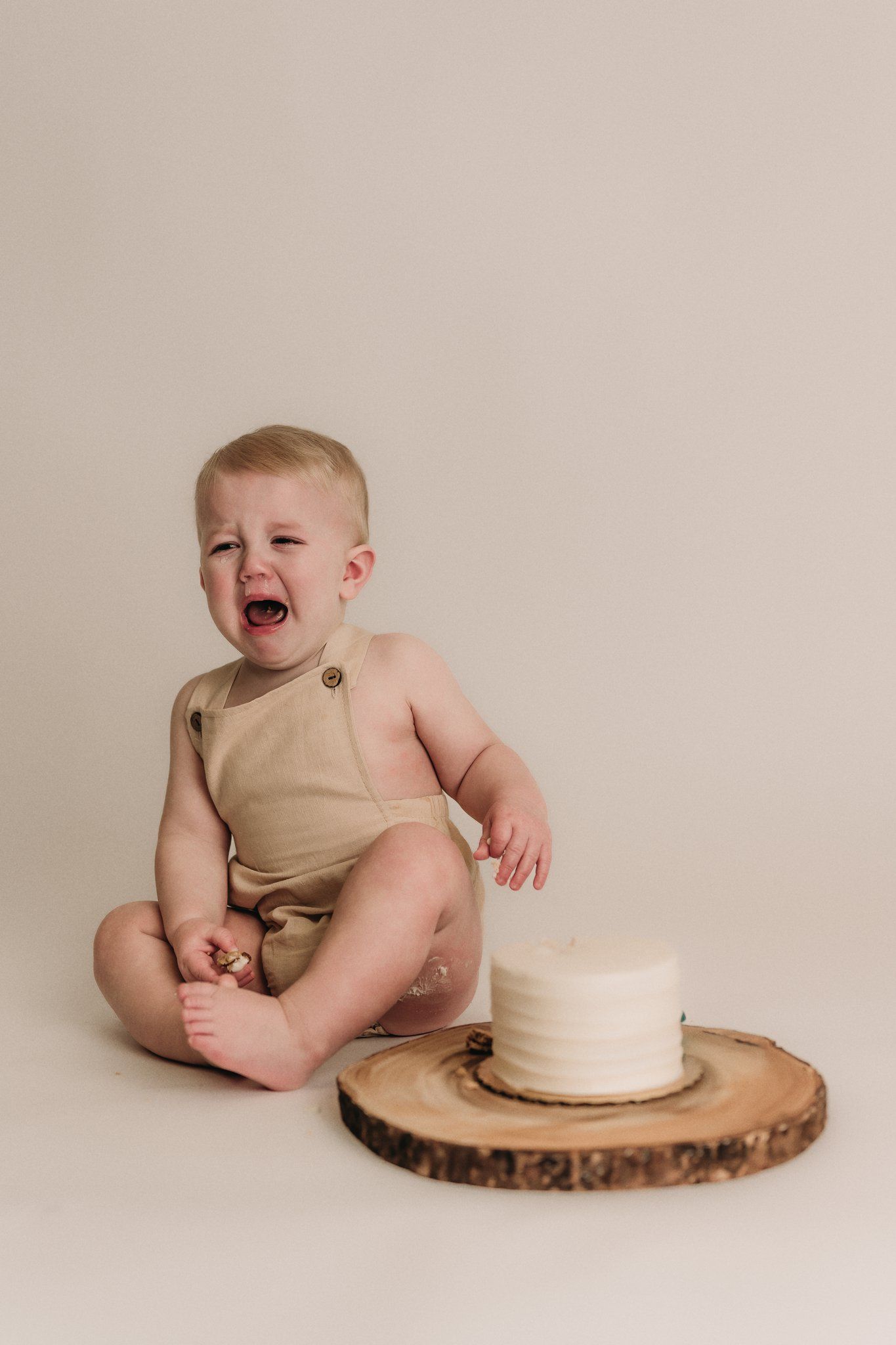 Baby in beige outfit expressing emotion next to white cake on wooden slice.