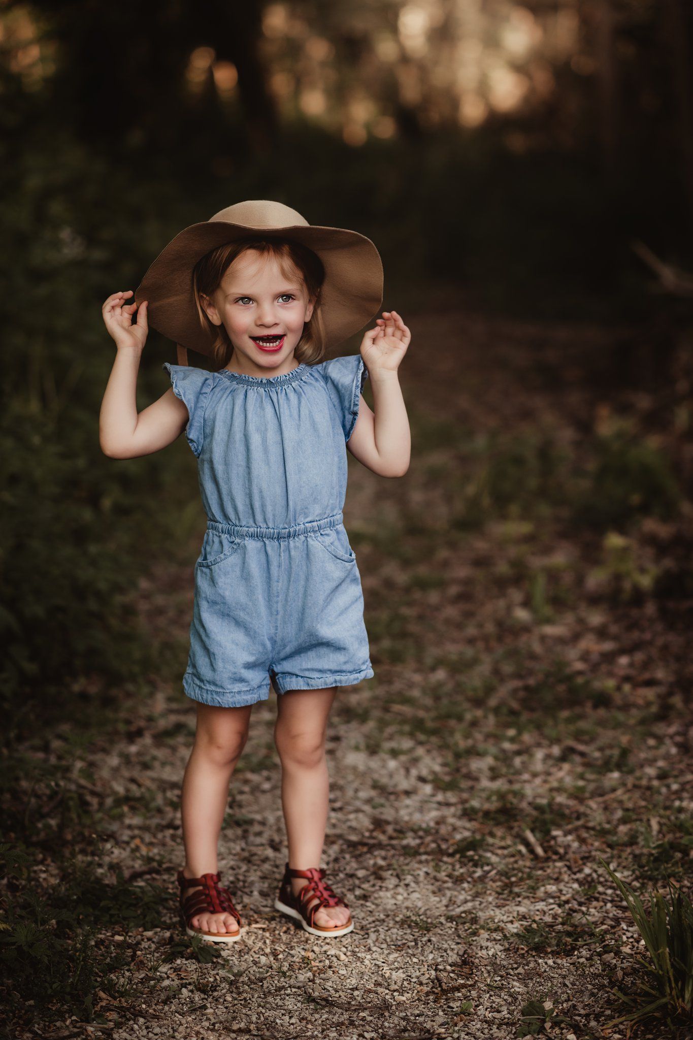 Child wearing denim romper and wide-brim hat standing on forest path.