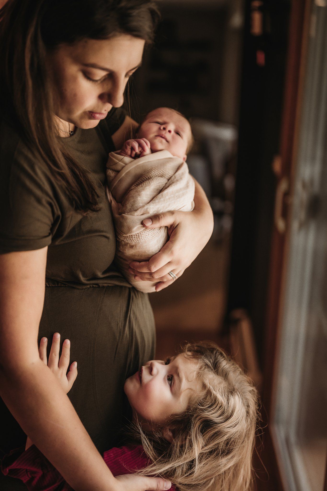 Tender family portrait with baby in beige blanket and child in red dress by window light.