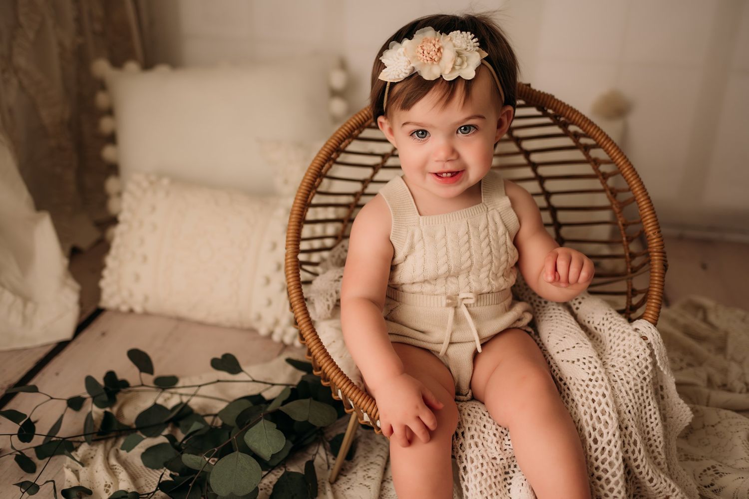 Baby poses in vintage-style cream romper on rattan chair with decorative greenery.