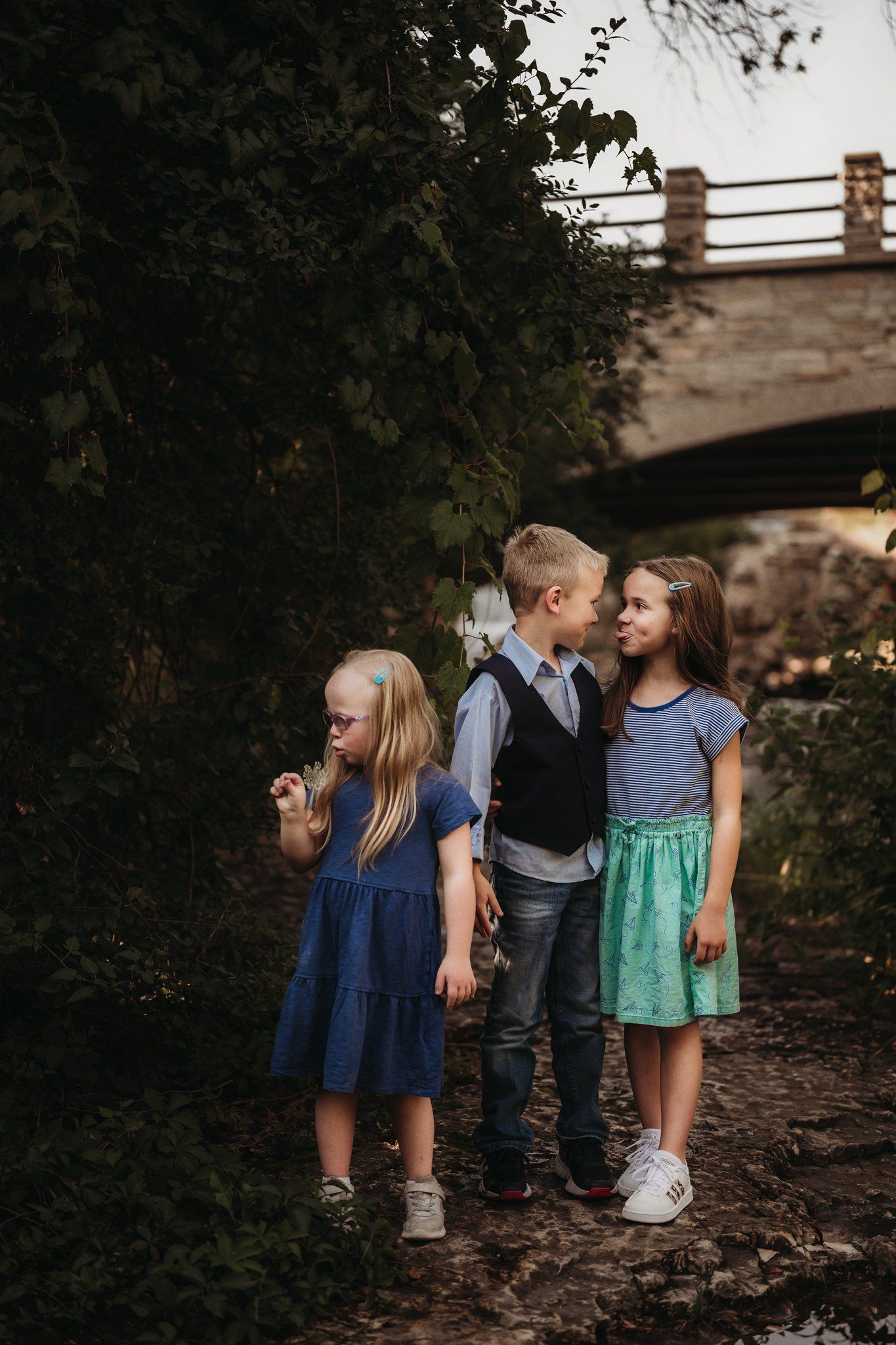 Children in casual attire pose together on a wooded path beneath a bridge.