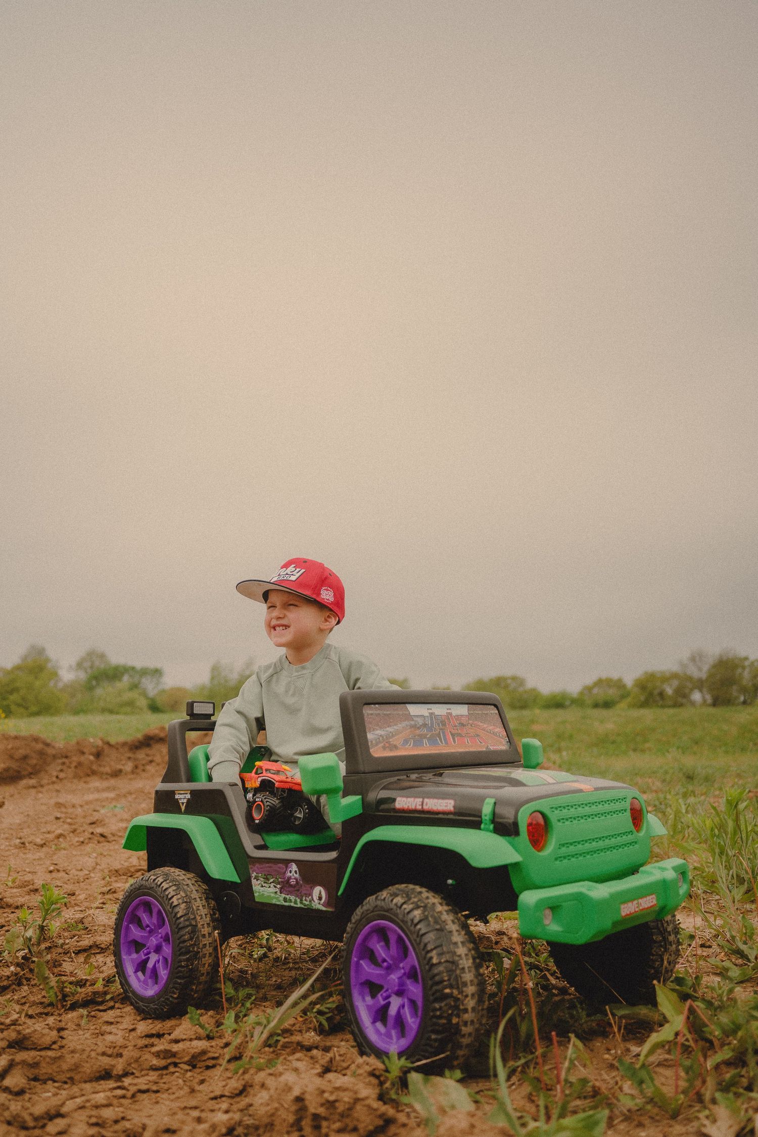 Young boy in red cap rides green Monster Jam toy Jeep on dirt field outdoors.