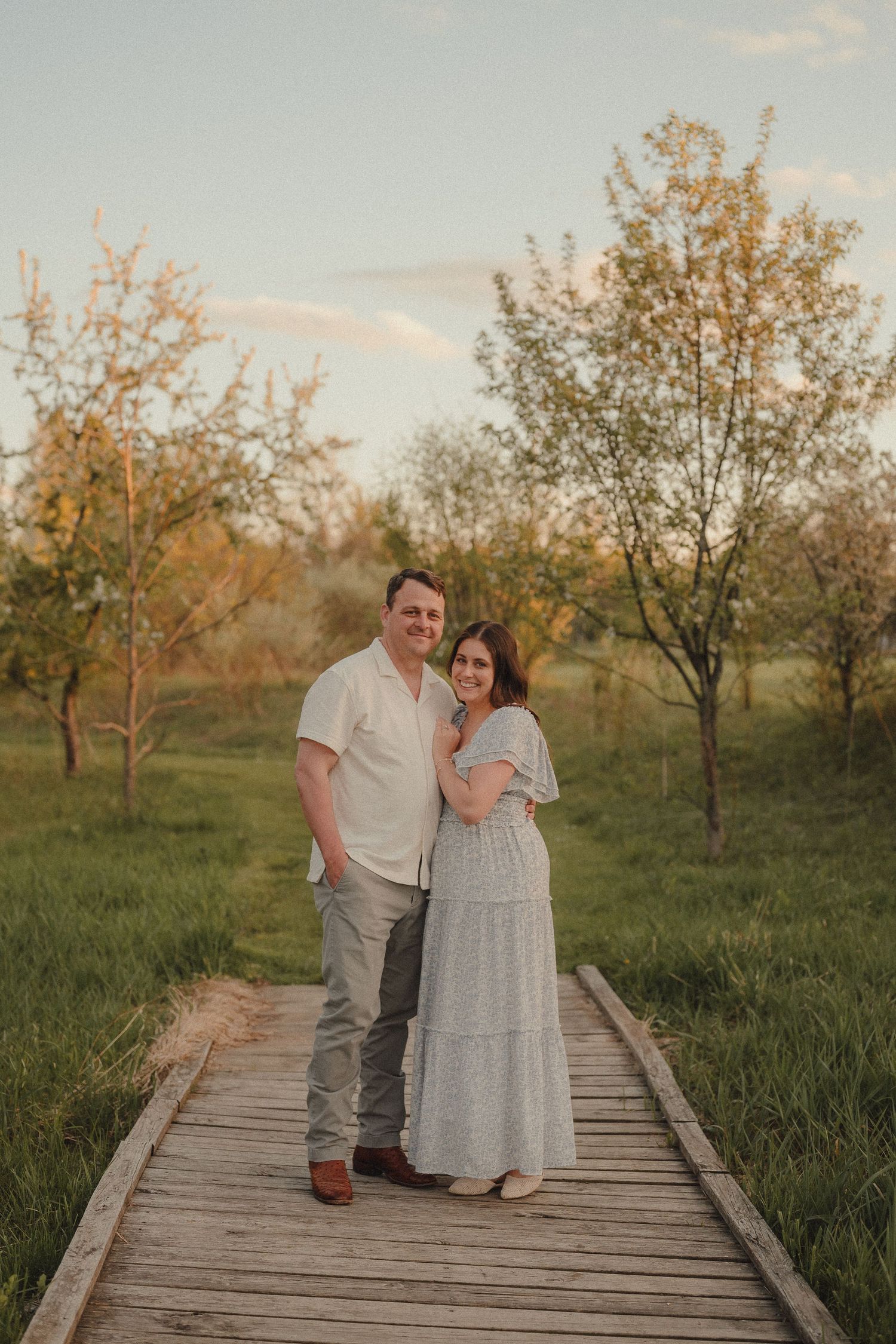 Couple standing on wooden boardwalk path surrounded by blooming trees at golden hour sunset.