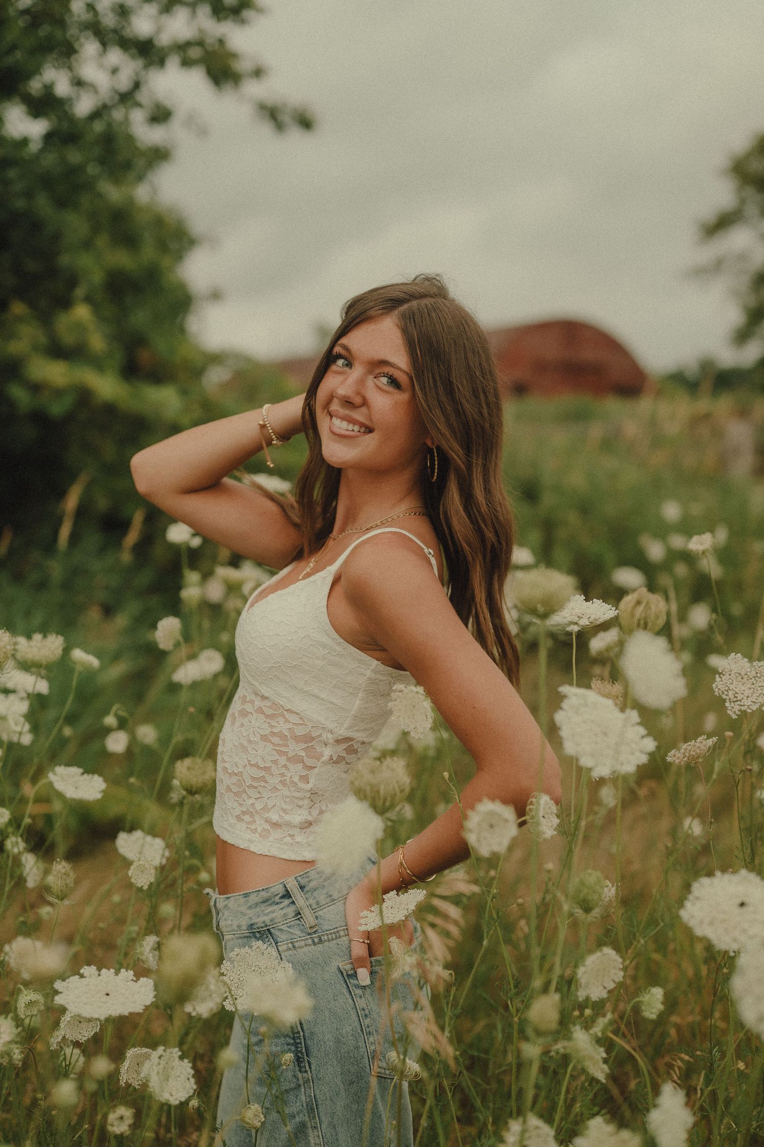 Smiling young woman in white lace crop top and jeans posing among white wildflowers in a field.
