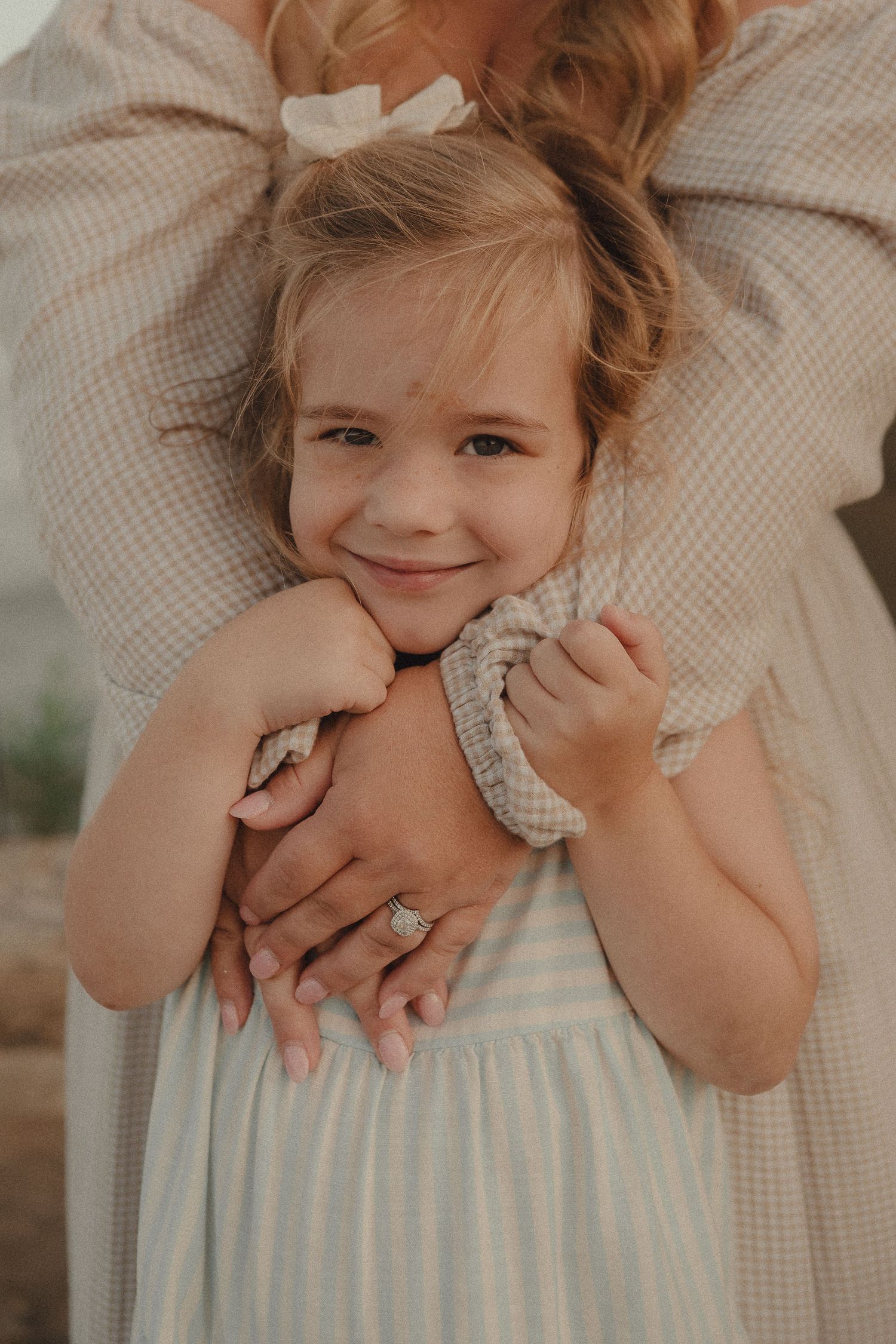 Smiling young girl in striped dress being hugged from behind by mother wearing diamond engagement ring.