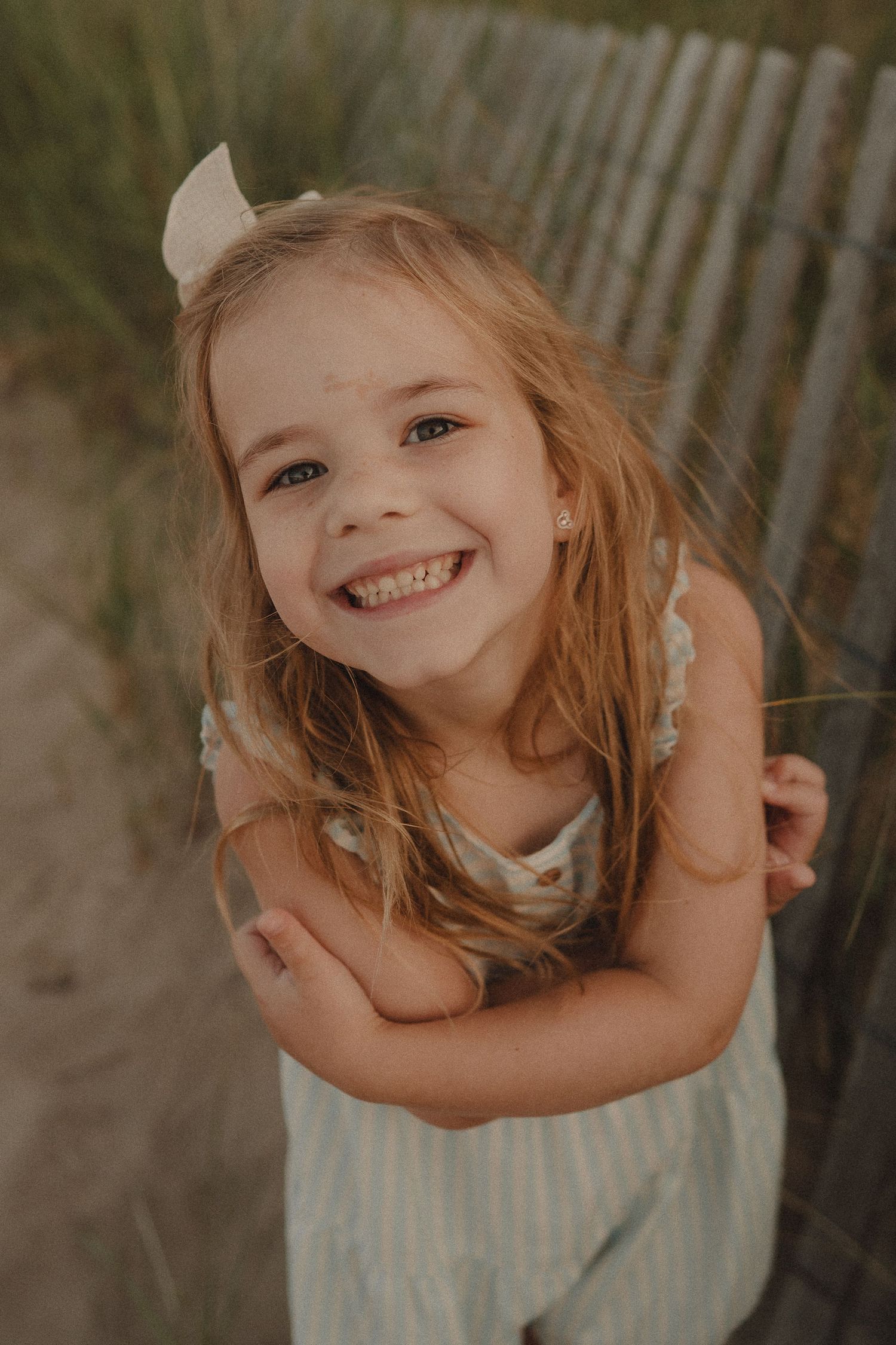 Smiling young girl with blonde hair and bow at beach fence.
