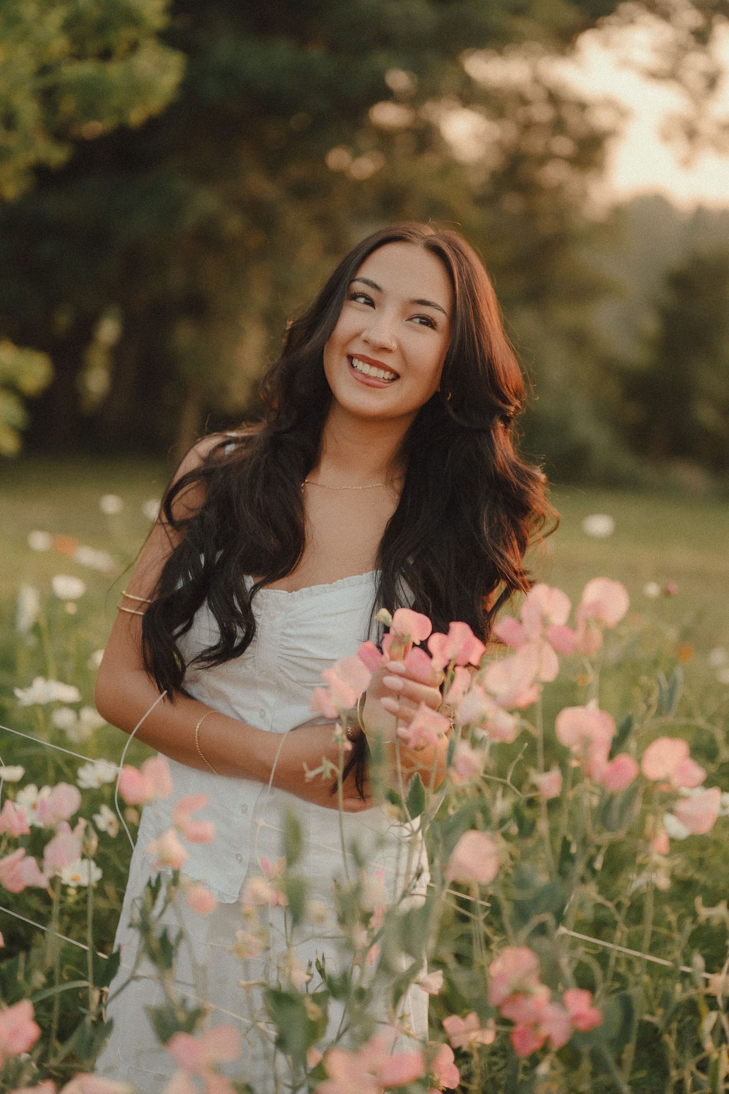Smiling woman with long dark hair holding pink flowers in a sunlit garden wearing a white dress.