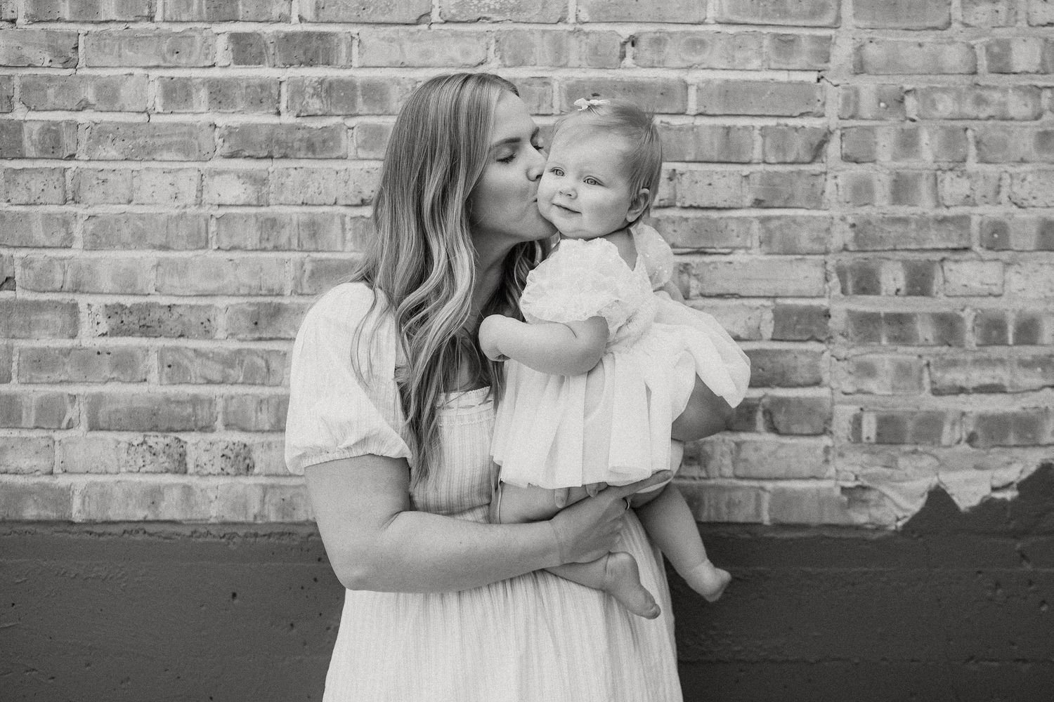 Black and white photo of a mother kissing her baby girl against a brick wall backdrop.