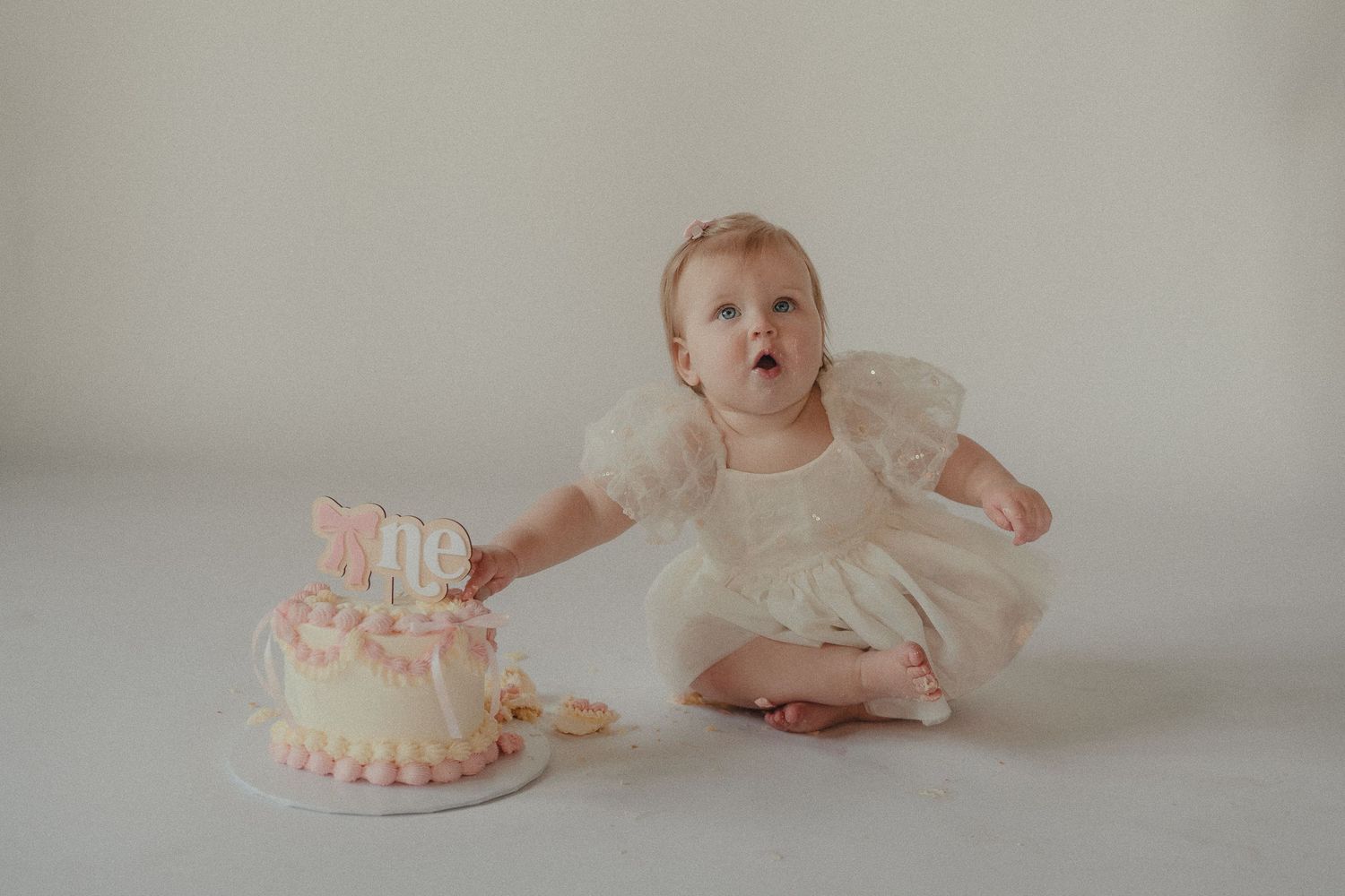Baby girl in white puff sleeve dress reaches toward her pink smash cake during her first birthday photo session.