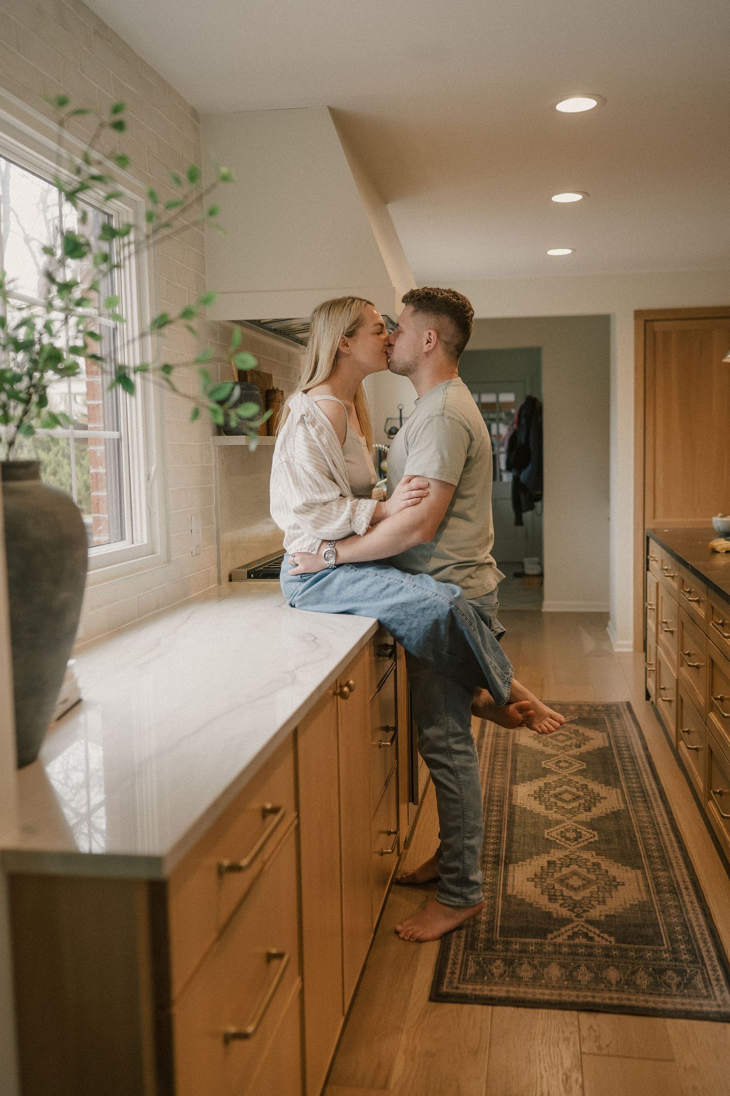 Couple sharing a romantic kiss on a white marble kitchen counter by a sunny window.