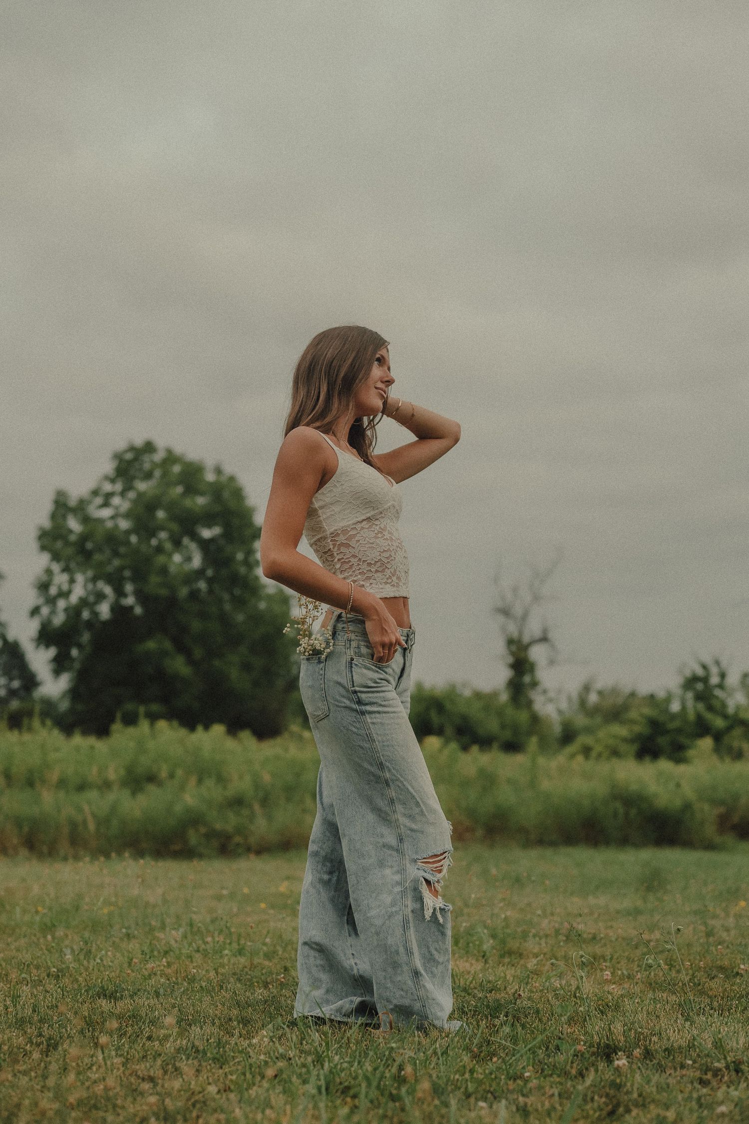 Young woman in white lace top and wide-leg ripped jeans standing in a green field under cloudy skies.