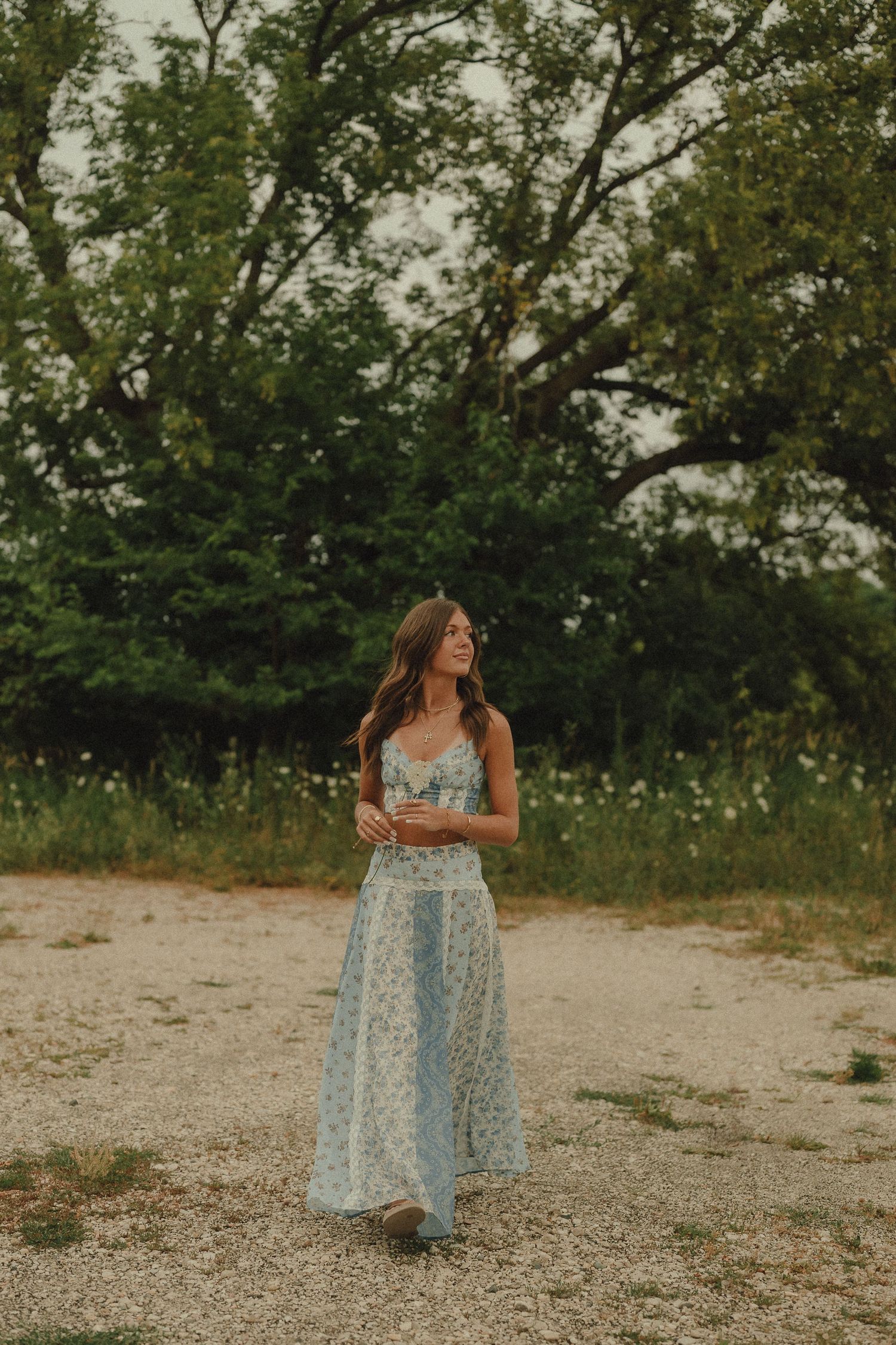 Young woman in light blue floral maxi dress walking on gravel path surrounded by green trees.