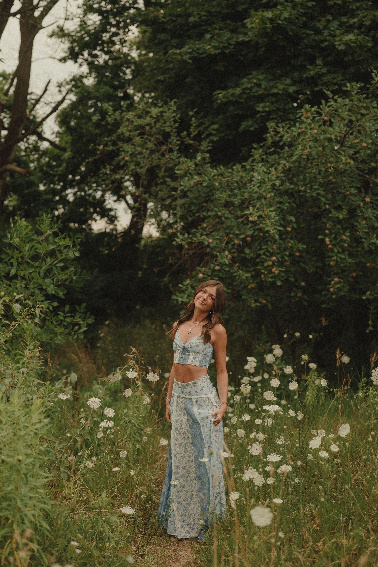 Young woman in blue floral crop top and maxi skirt smiling in a wildflower meadow surrounded by lush green trees.