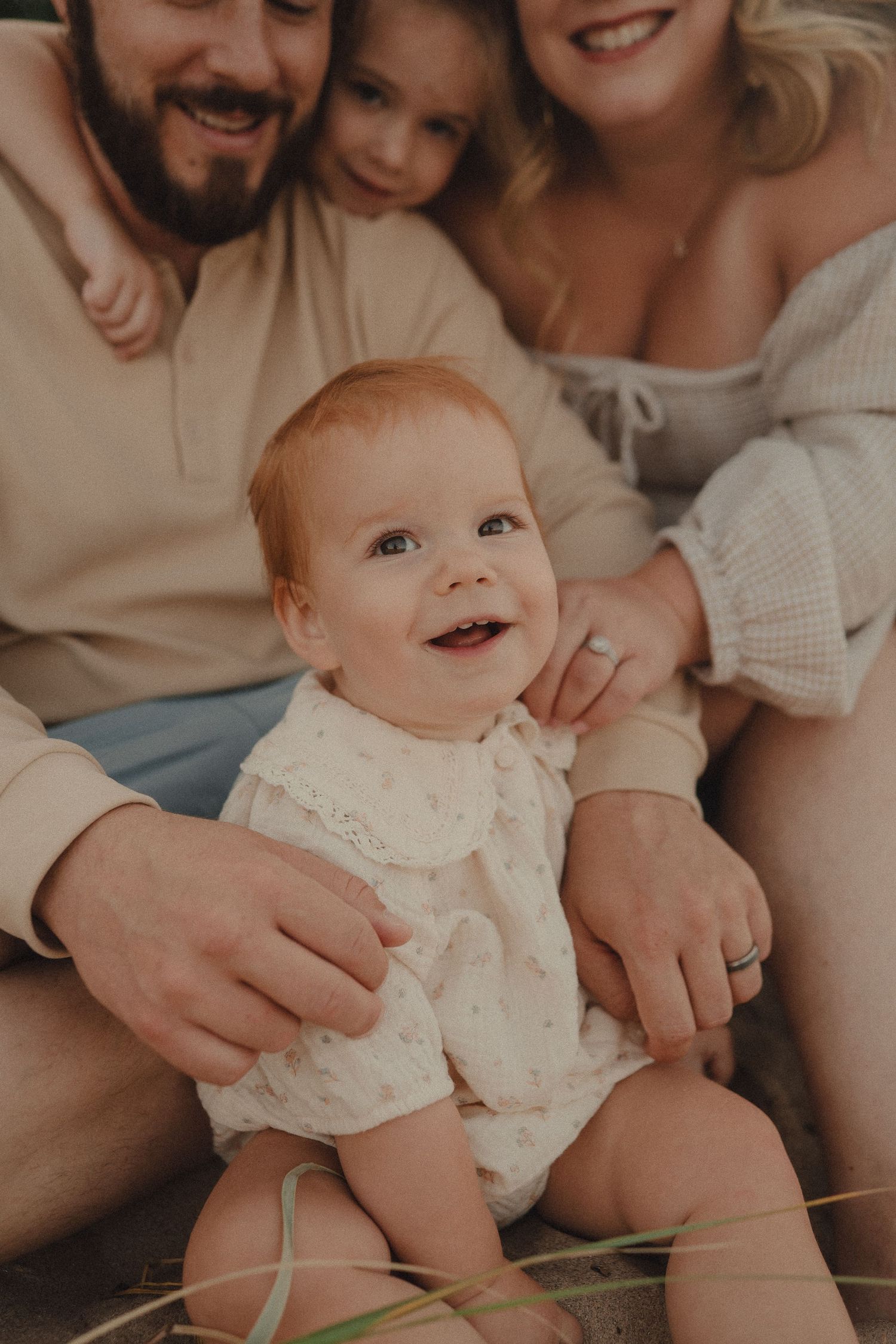 Happy baby in floral romper smiling at camera while family of four sits together on sandy beach.