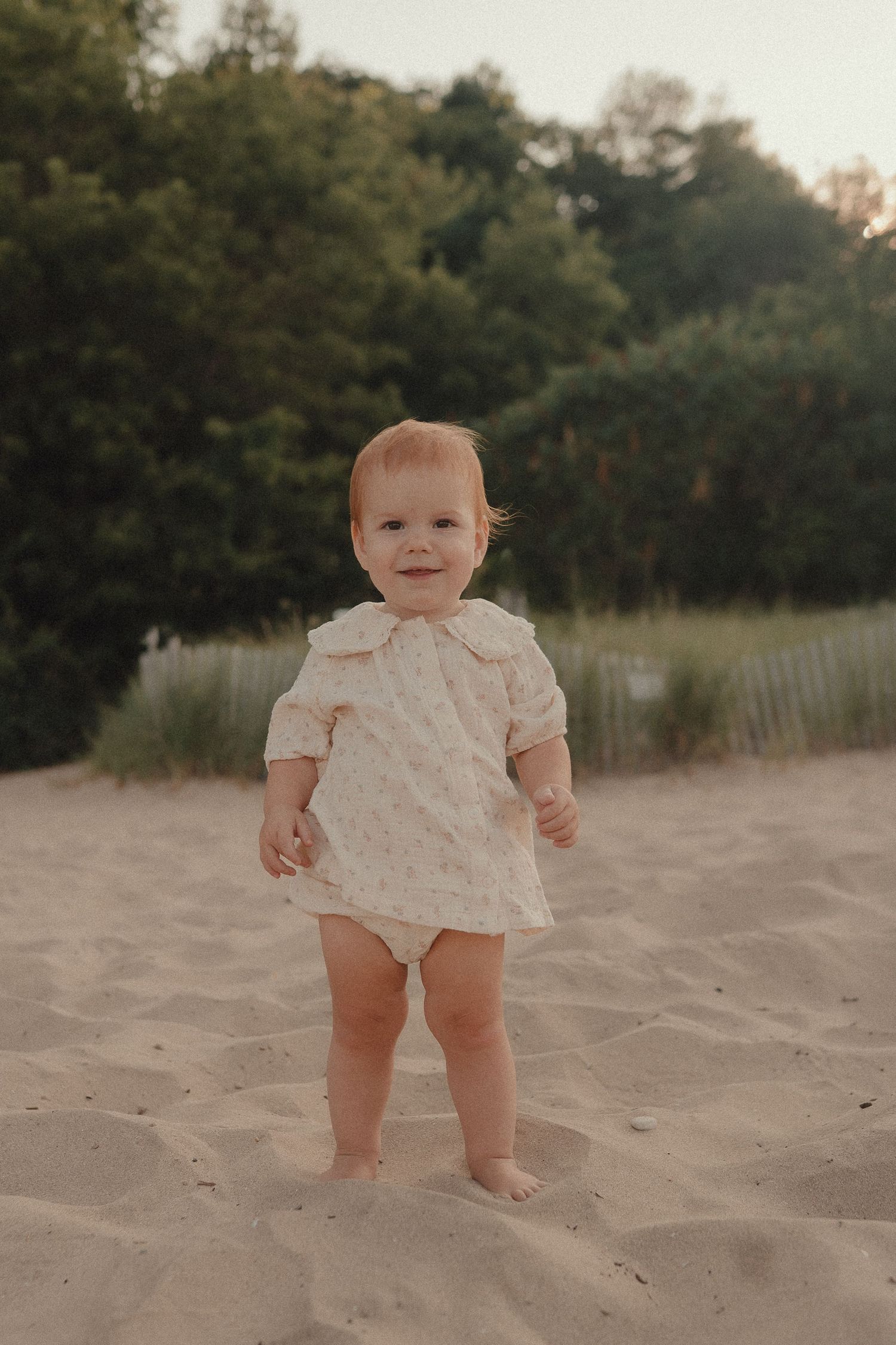 A smiling baby girl in a floral dress stands barefoot on sandy beach dunes with green trees in the background.
