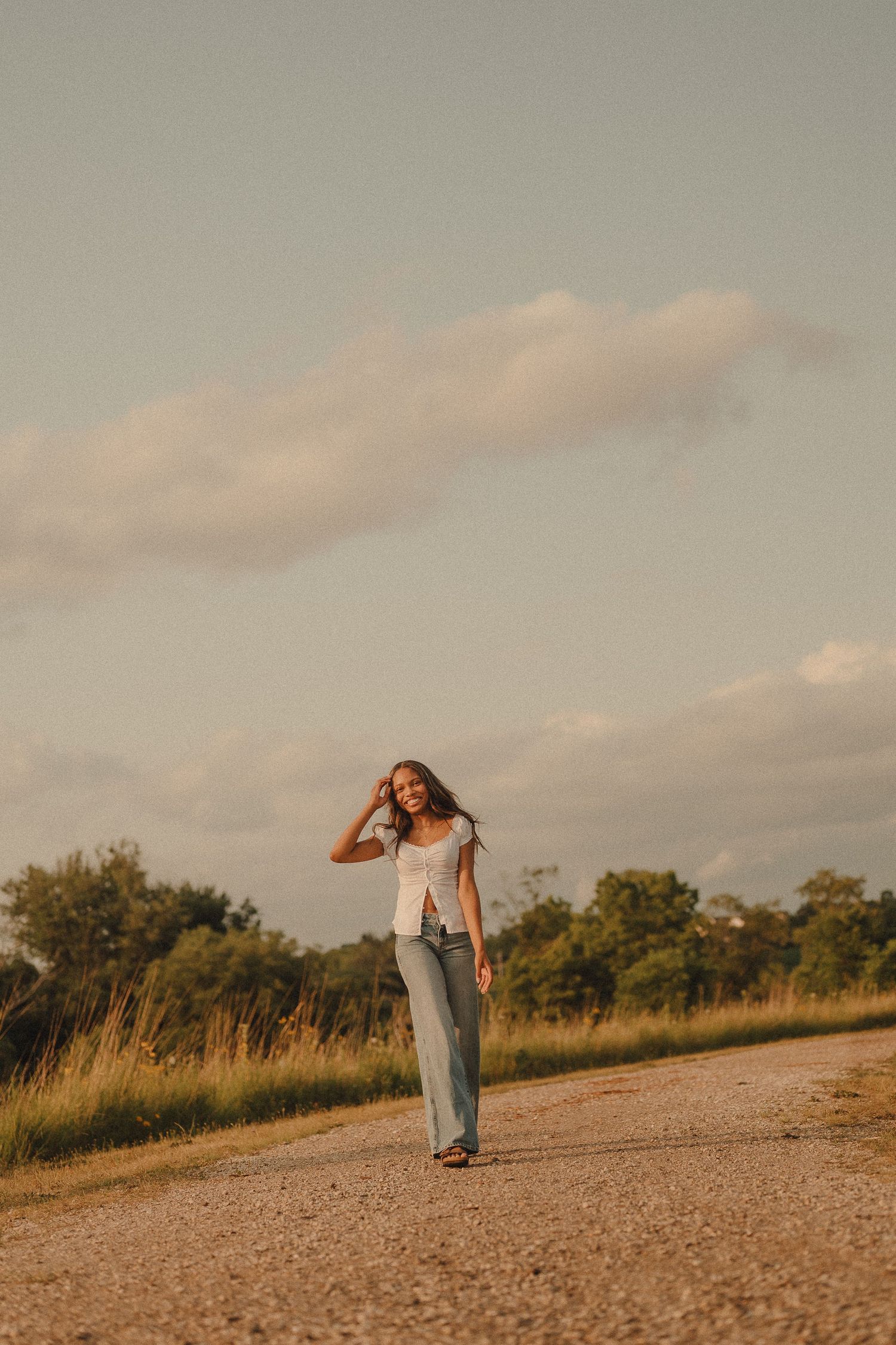 Young woman in white top and jeans walks along a rural dirt road at golden hour with trees in background.
