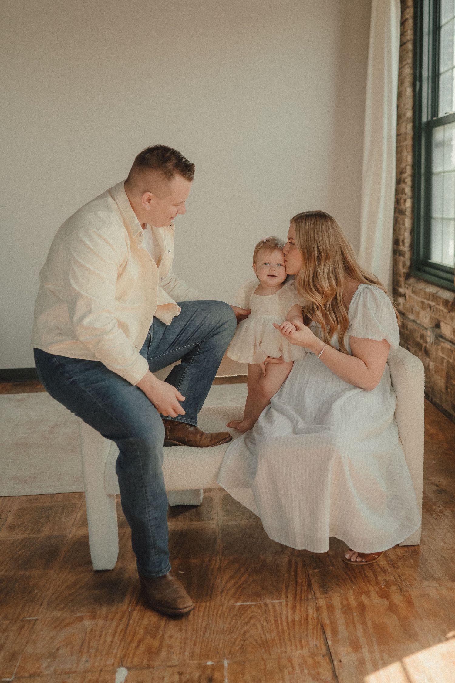 A young family with a toddler shares a joyful moment on a white chair in a bright, warmly lit studio setting.