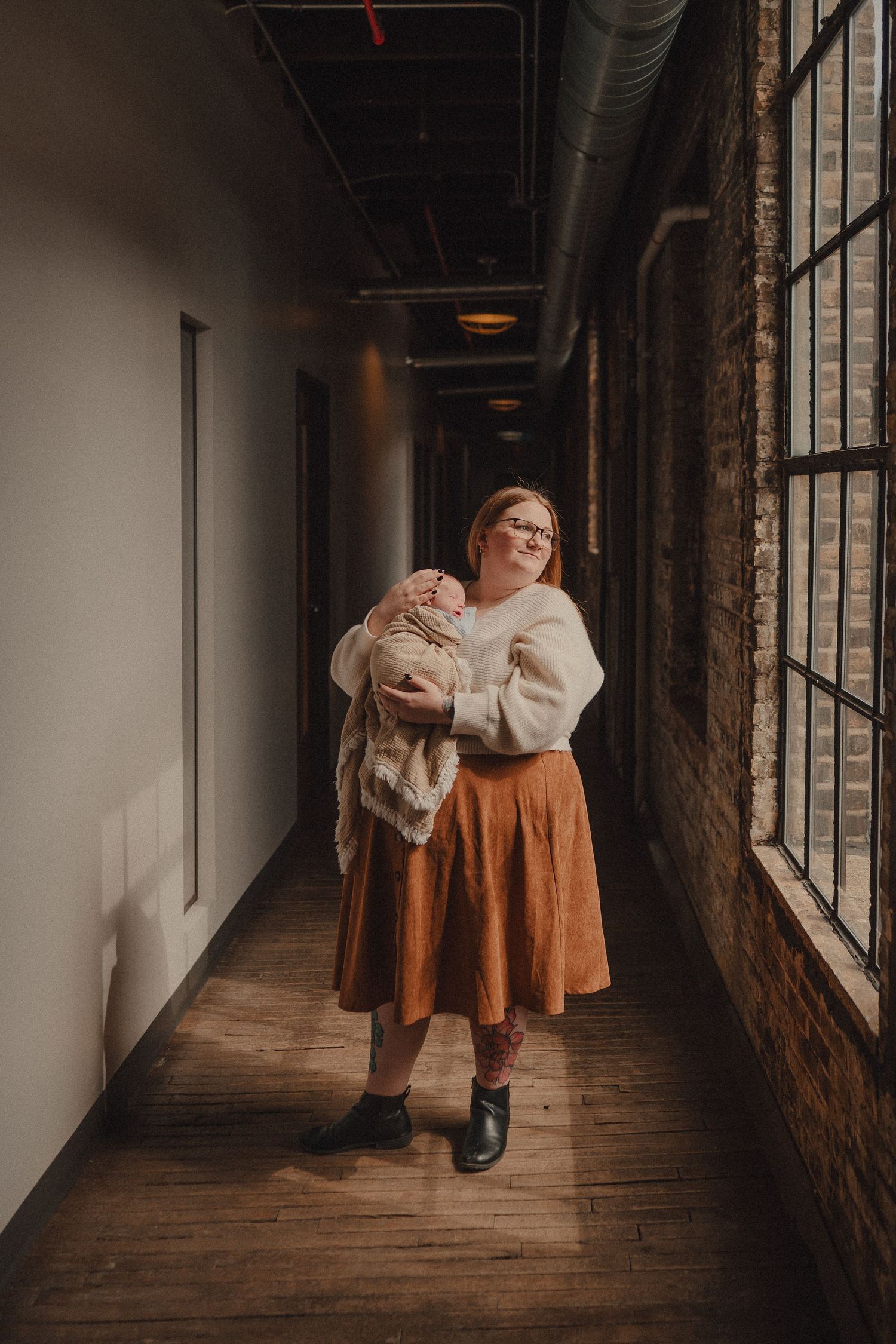 A redheaded woman in a cream sweater and rust skirt holds a newborn baby in a sunlit brick hallway.