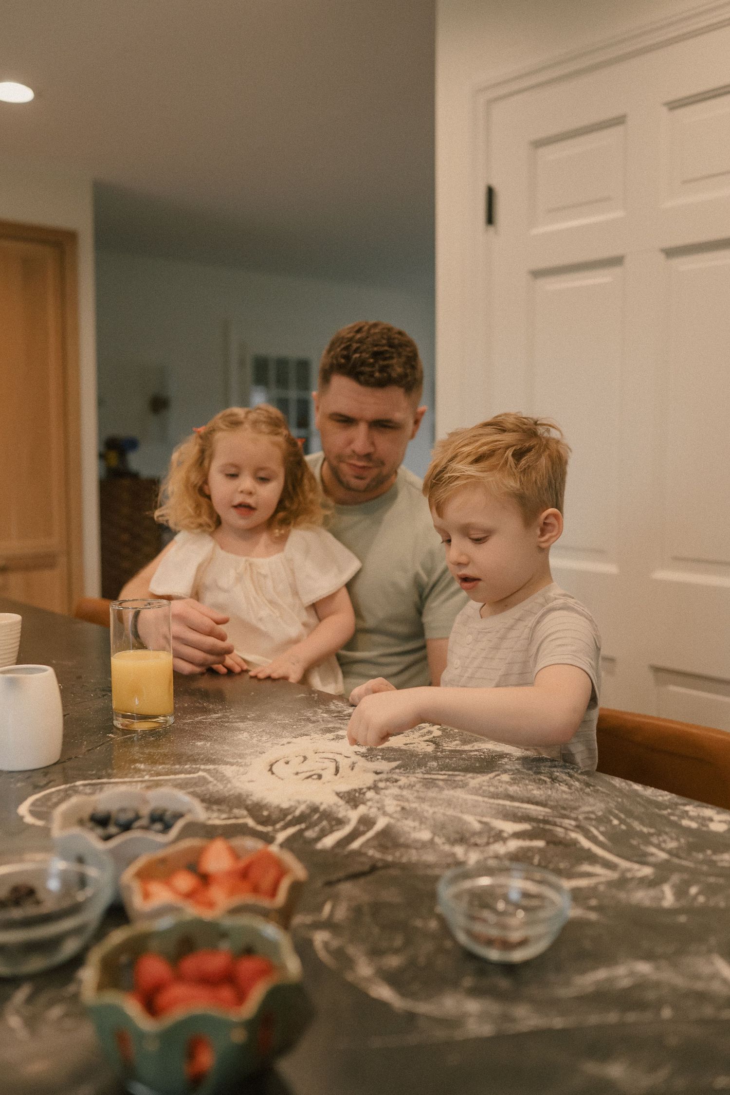 Father and two young children playing with flour on dark kitchen counter.