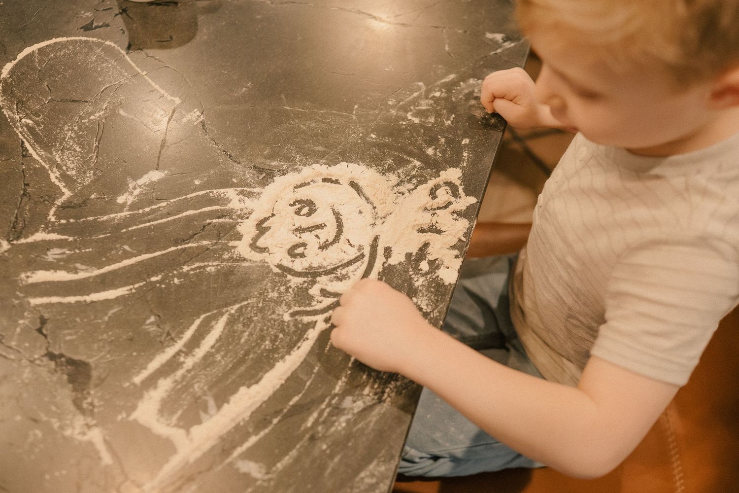 Young child drawing a figure in white flour spread on a dark table surface.