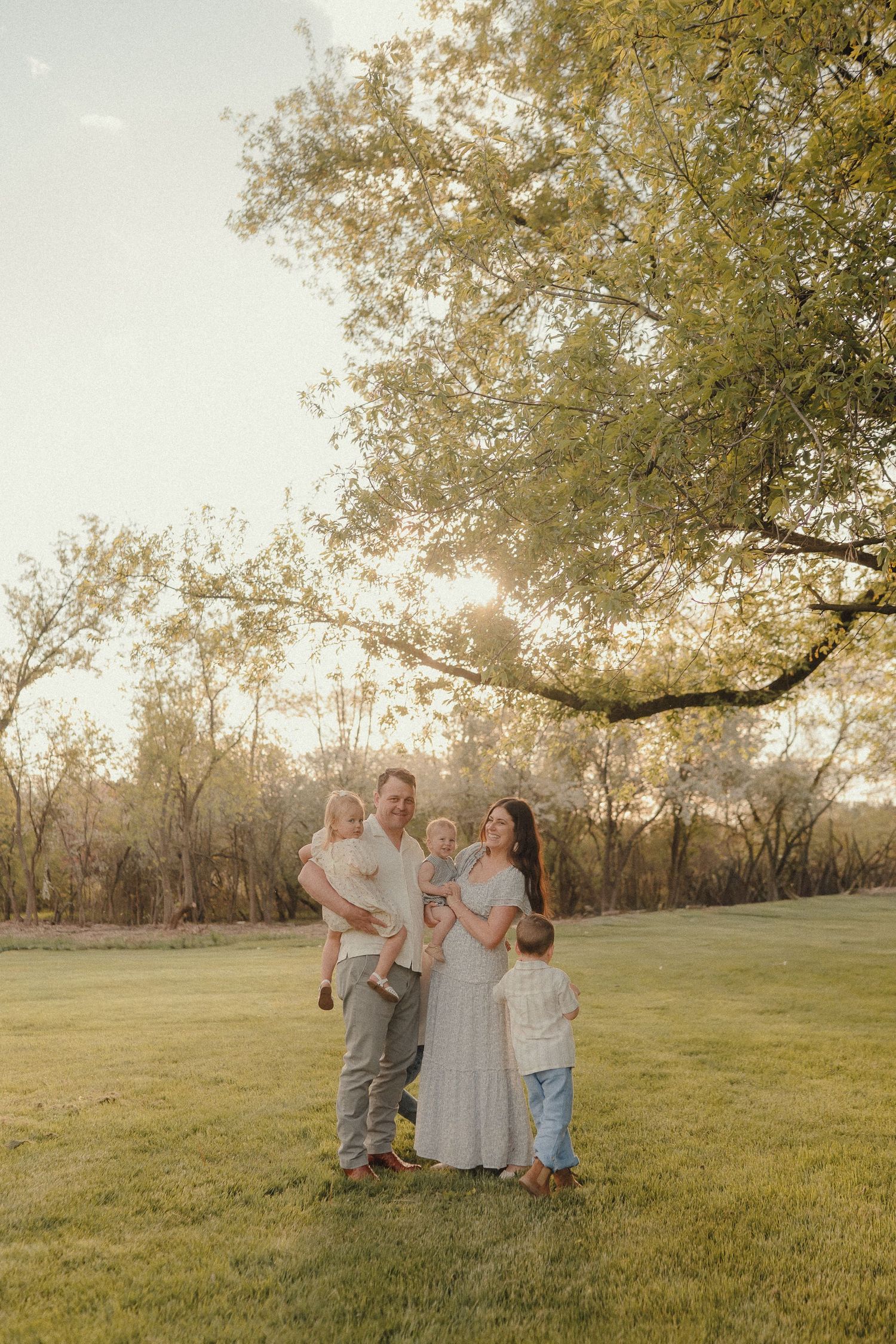 Happy family of five posing together on green lawn at sunset, parents holding young children near a large leafy tree.