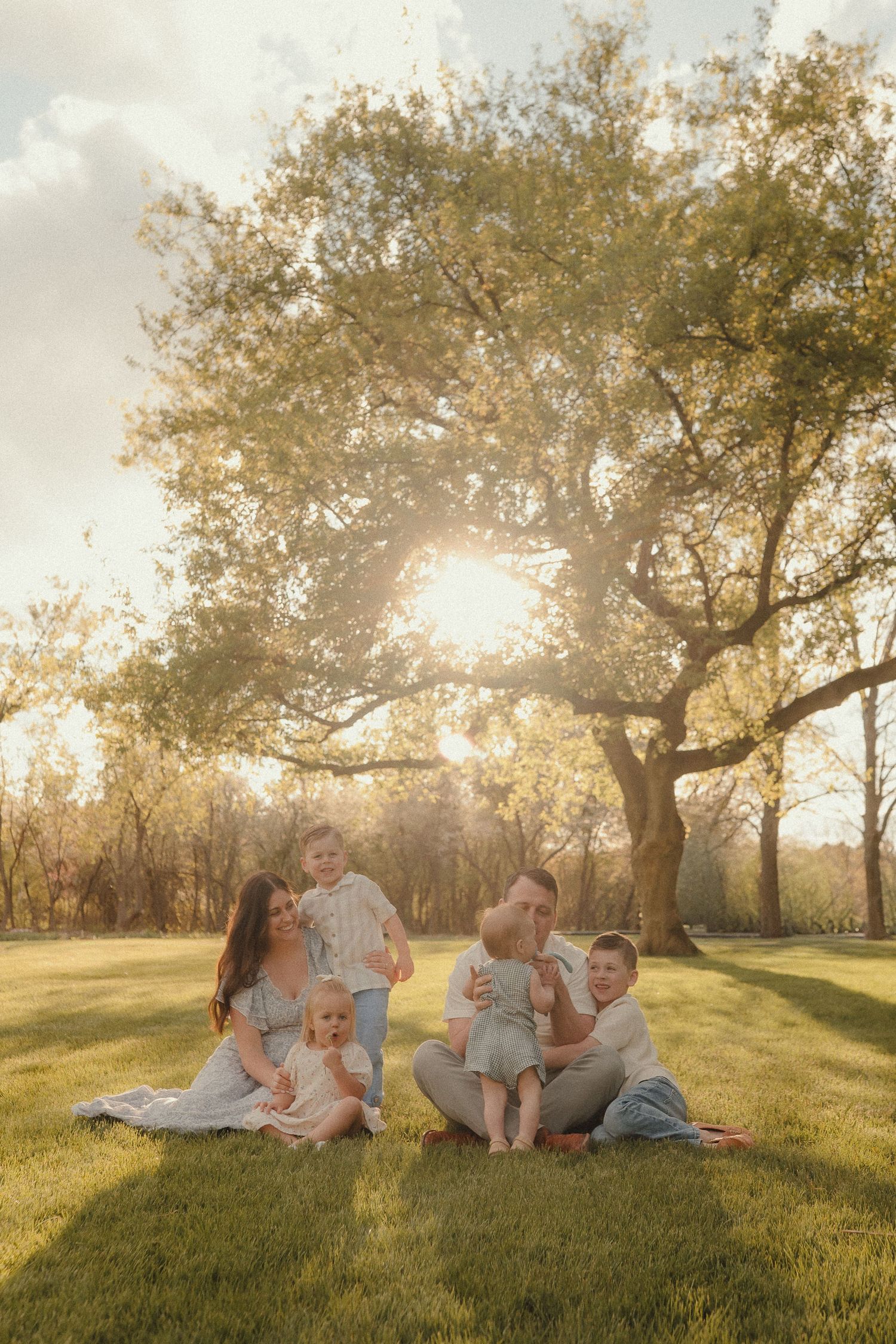 Family of six sitting on grass at golden hour beneath a large tree, parents holding young children in warm sunlight.