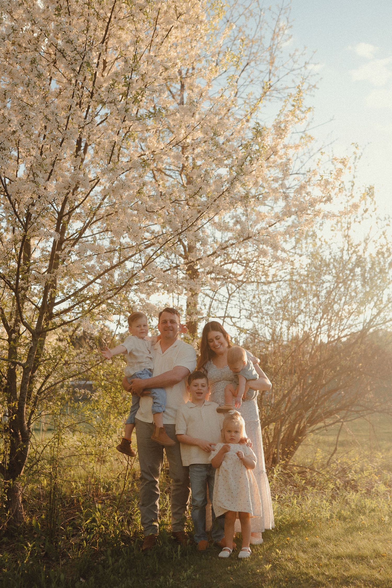 Happy family with three children standing together under flowering white tree during warm golden hour sunset.