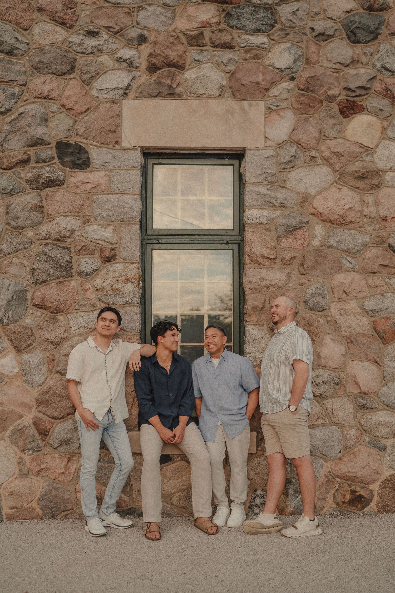 Four men posing casually against a rustic stone wall with a window backdrop.