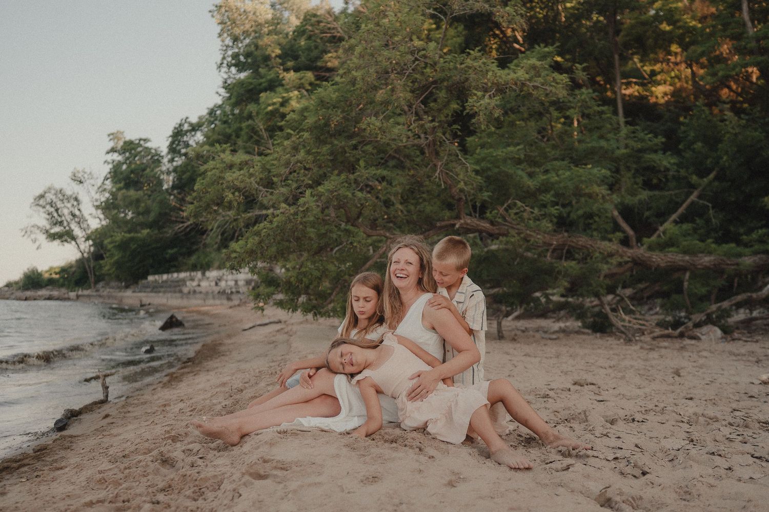 Family of four sitting together on a sandy beach, smiling and hugging near the shoreline with lush green trees behind.