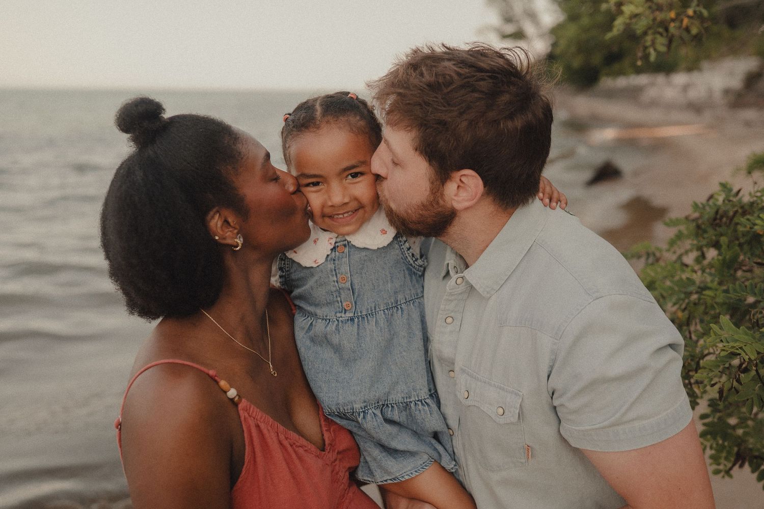A loving interracial family shares kisses on their smiling young daughter's cheeks at a scenic beach.