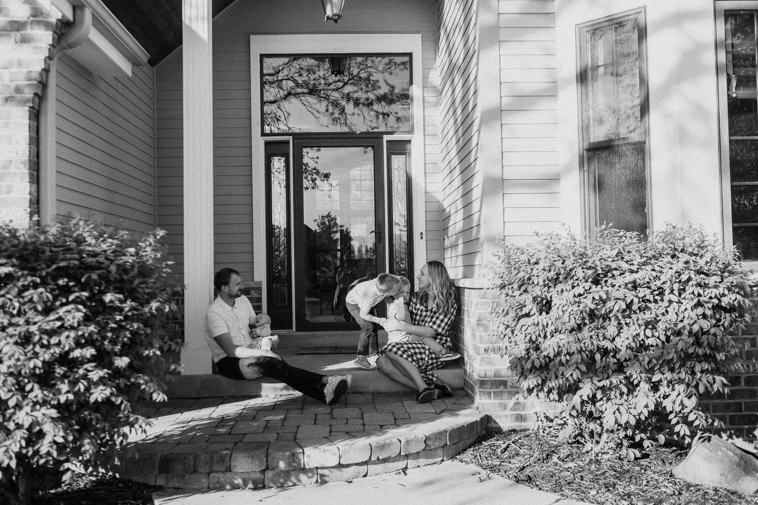 Family relaxing together on the front porch steps of a suburban home in a candid black and white photo.