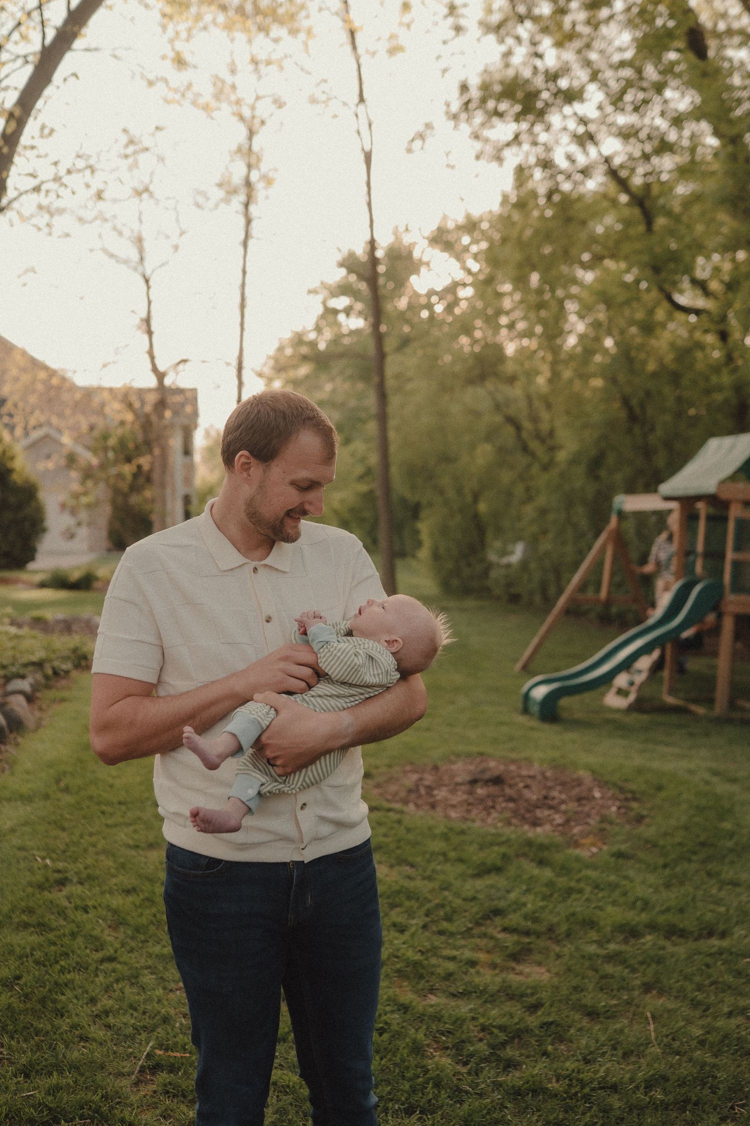 Smiling father in beige polo holds newborn baby in backyard with playground equipment behind him.