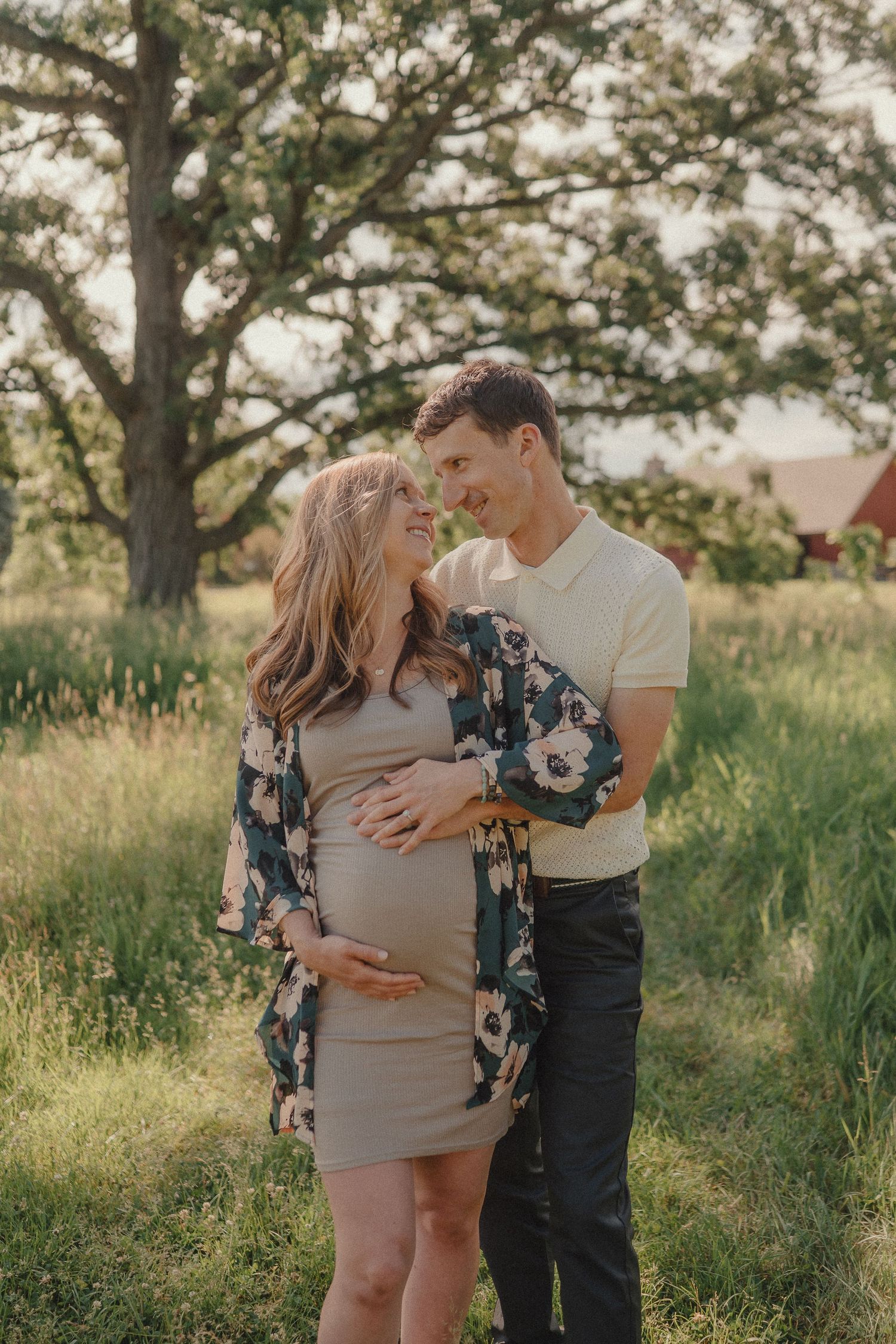 A smiling pregnant couple embraces tenderly in a sunlit green field with a large oak tree and red barn behind them.