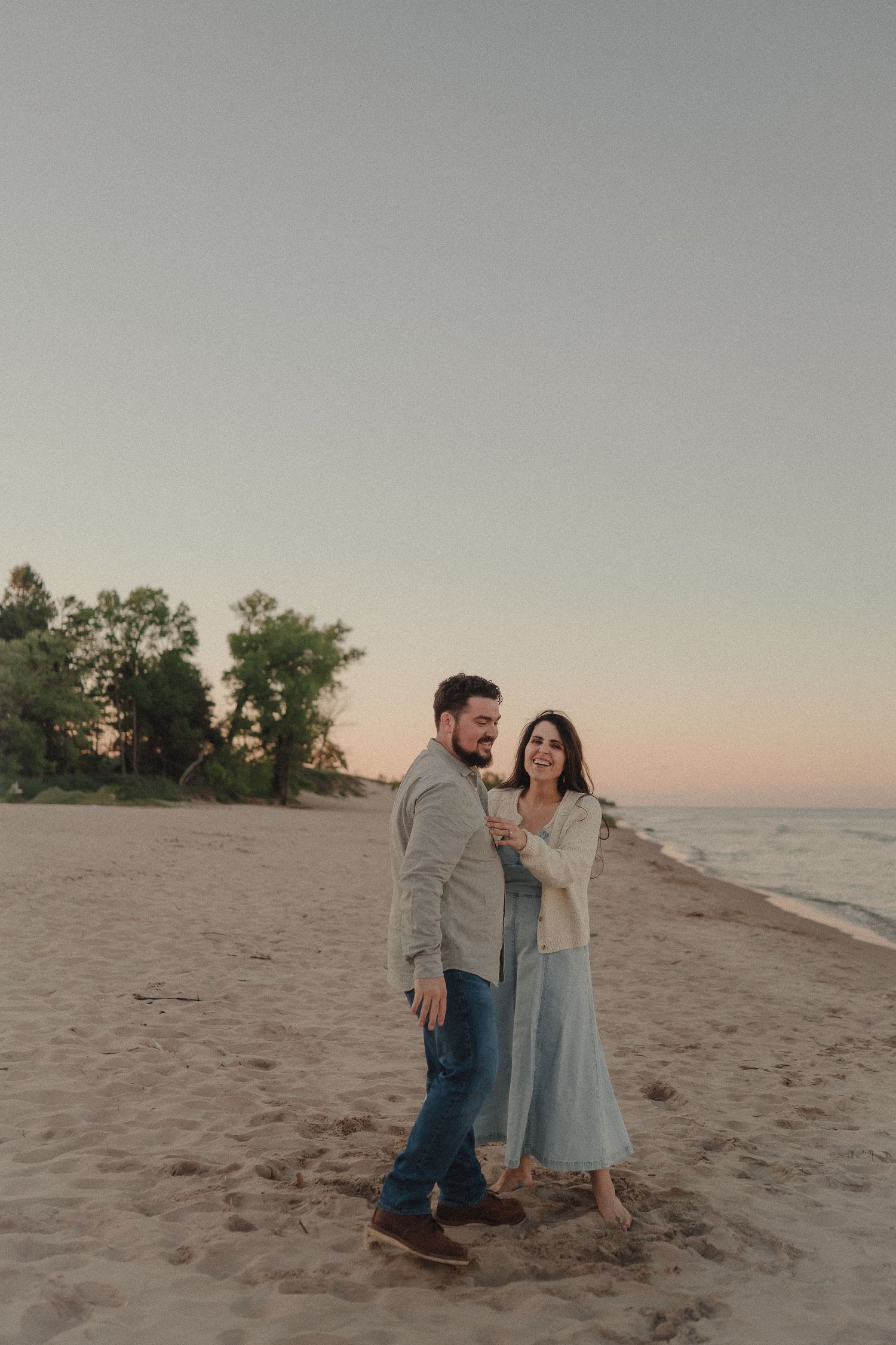 Happy couple laughing and walking together on sandy beach at golden hour near ocean shore.