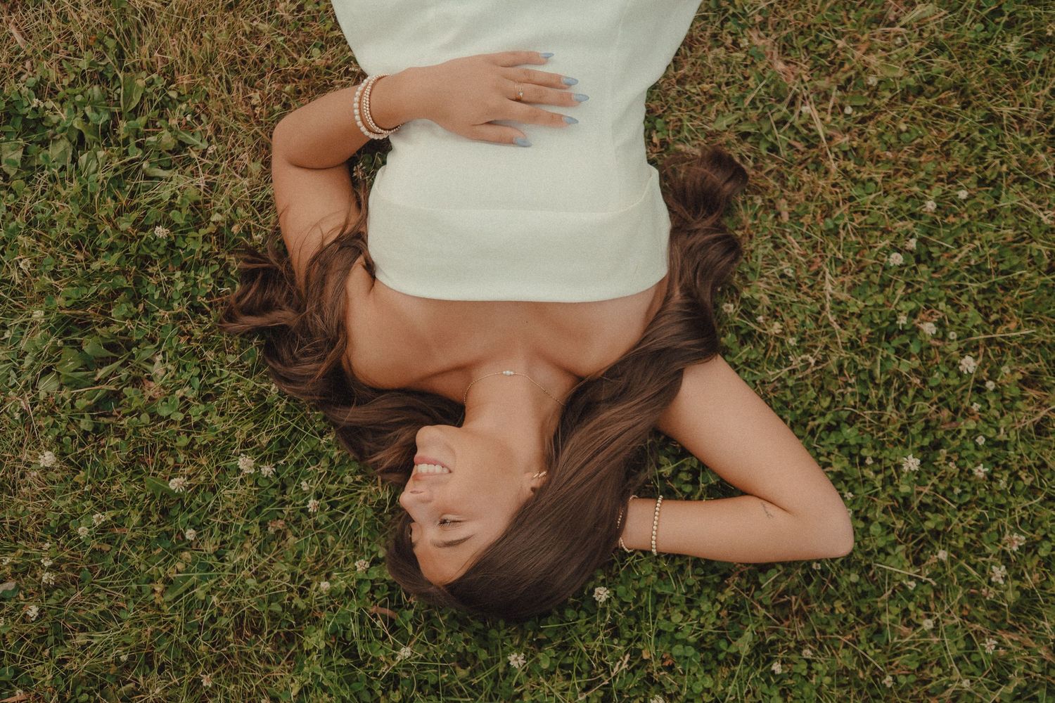 Young woman in white dress lying on green grass with long brown hair spread out, viewed from above.