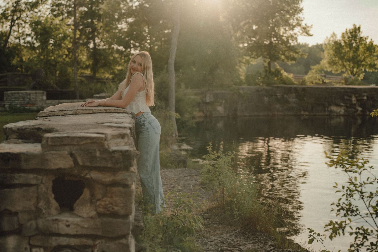 A blonde woman in light blue jeans leans on a stone wall by a serene pond at golden hour.
